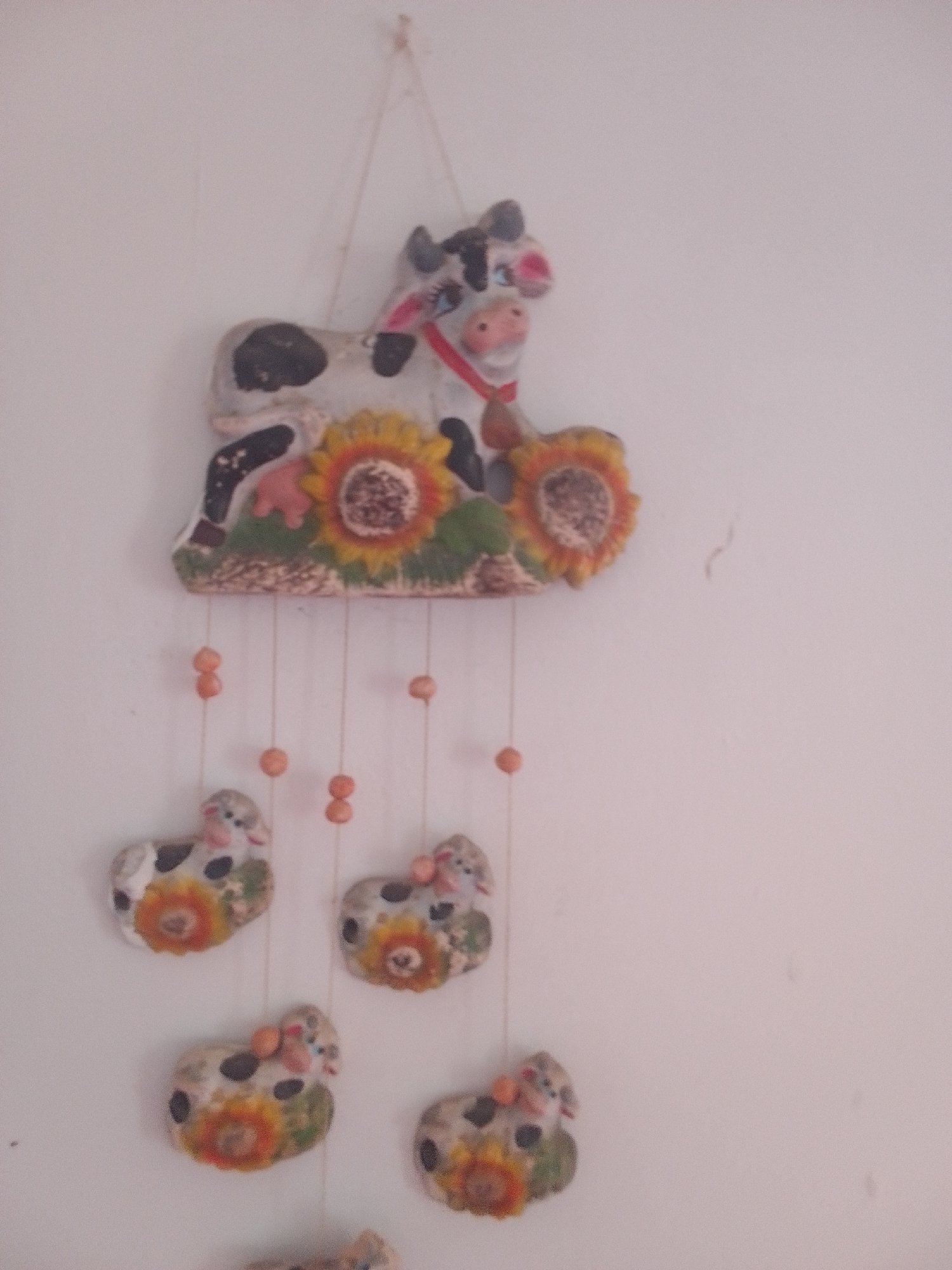 This media file is a photograph of a decorative wall hanging. The main subject is a ceramic decoration featuring a cow with sunflowers. The cow is primarily white with black spots, a pink nose, and a red ribbon around its neck. It sits atop a base decorated with two sunflowers, one on its left and one on its right.
Below the main cow figure, several smaller, similar ceramic cow figures hang at intervals on strings. Each of these smaller cows also has black spots, pink noses, and is adorned with a sunflower. Small, round, orange beads are interspersed along the strings holding the lower cow figures.
The background is a plain, light-colored wall, suggesting an indoor setting. There is no visible text in the image. The lighting appears to be natural, casting a soft illumination on the decorative pieces. The overall scene is simple and domestic, likely a home decor item. The image quality is slightly blurred, making precise details hard to discern. The location context of La Tagua, Venezuela, suggests this might be a handicraft or souvenir from that region.