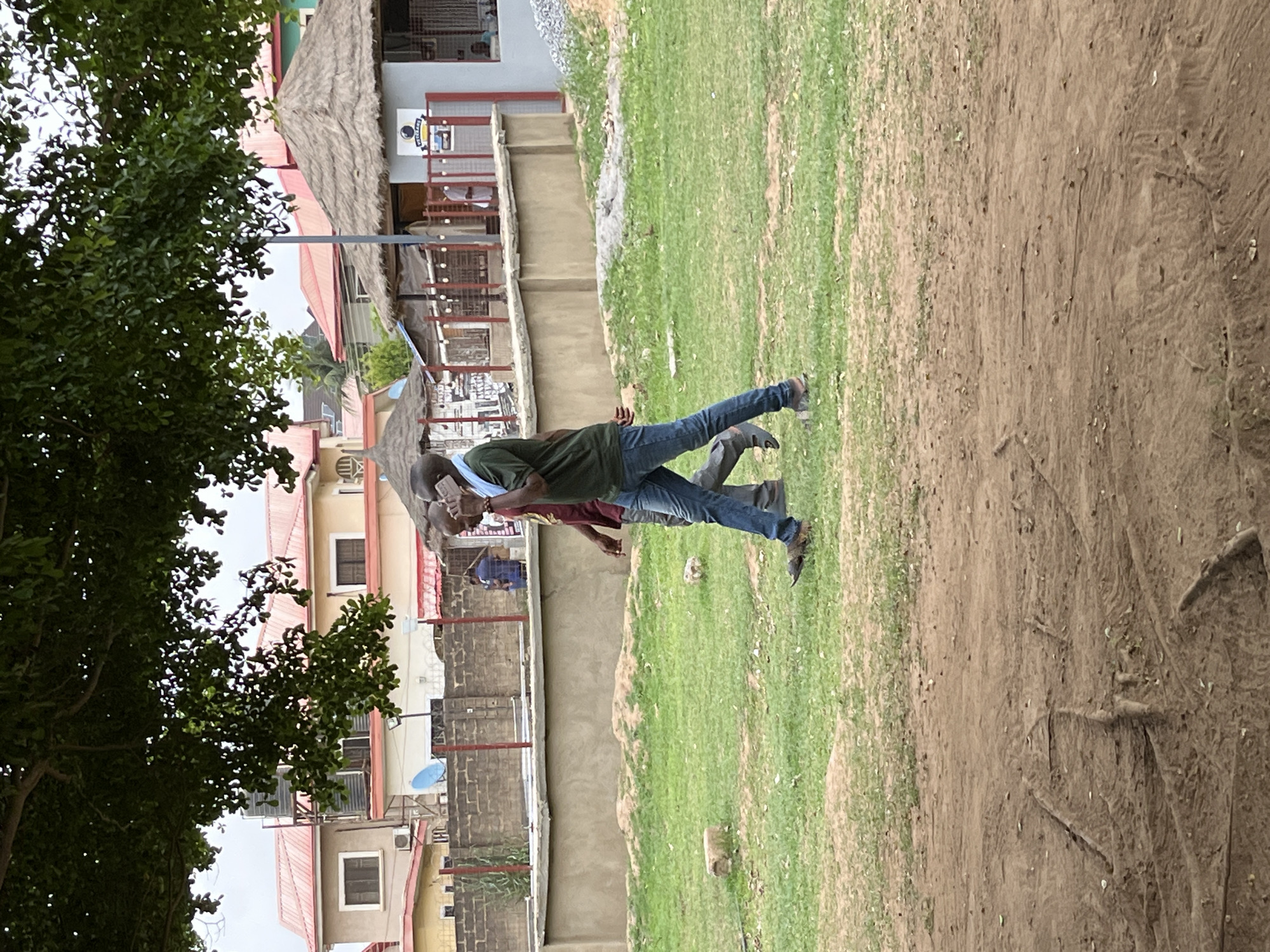 The image shows two people walking in Galadimawa, Nigeria. One person is wearing a green shirt and jeans, and the other is wearing a maroon shirt. They are walking on a grassy area next to a dirt path. Buildings are visible in the background, as is a tree.