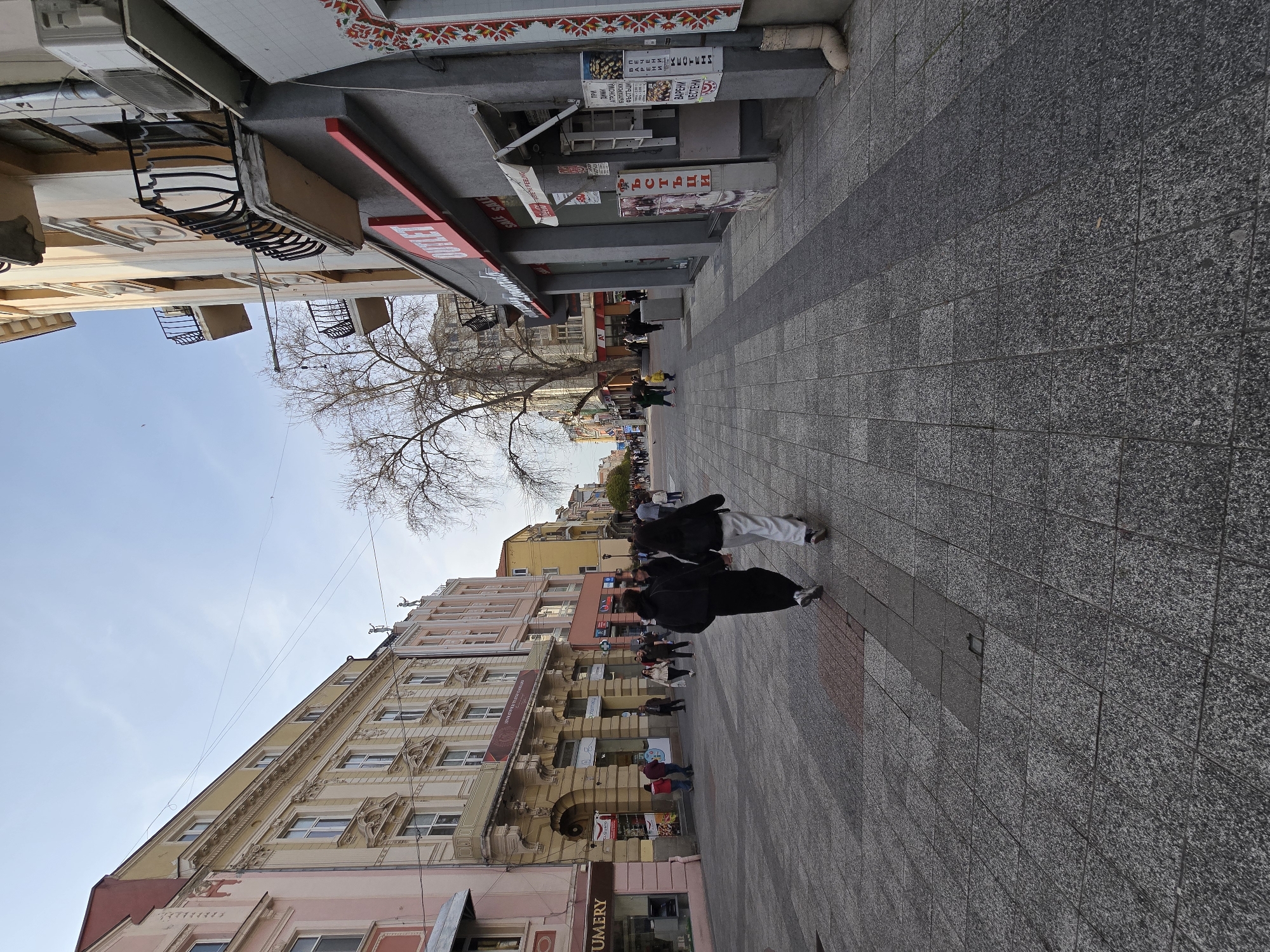 Here's a description of the image:
The image presents a street scene in a city of unknown location, during daylight. The scene is captured at street level, showing the sidewalk, the facades of buildings lining the street, and pedestrians moving along the sidewalk.
The buildings are of varying architectural styles and heights, with some exhibiting detailed ornamentation and balconies. There are signs and storefronts visible, suggesting a commercial area. A few of the signs have text in Cyrillic.
Pedestrians, most of whom are wearing casual attire, are present on the sidewalk. A bare tree extends its branches over the street, adding to the urban landscape. The pavement is composed of gray stones. The sky is light blue.