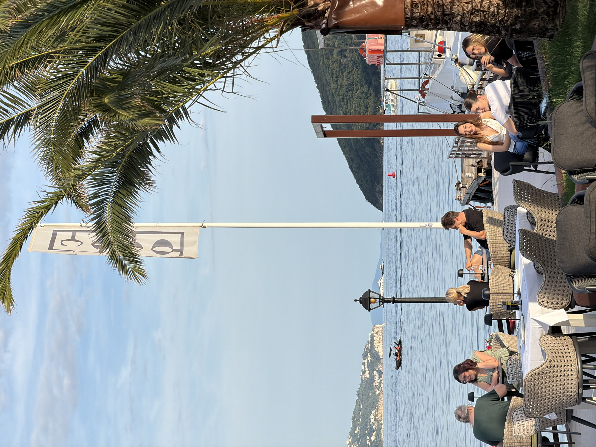 This image depicts a vibrant outdoor dining scene at a waterfront restaurant or cafe in Budva, Montenegro, during what appears to be late afternoon or early evening, characterized by warm, golden light.
In the foreground, a restaurant patio is furnished with numerous tables and chairs. Some tables are set with white tablecloths, dinnerware, and small, modern table lamps. The chairs are primarily made of woven rattan in light brown tones, though some darker, modern chairs are also visible. A large, green-leafed palm tree, partially framing the right side of the image, has its trunk wrapped in a brown material. Below the palm tree, a patch of green grass is visible.
Several groups of people are seated at the tables, engaged in conversation and relaxation. On the left side, an older man in a green t-shirt sits facing a woman in a light green patterned dress and glasses, both looking at each other across a table. Behind them, another woman with blonde hair in a black top is seated with a man in a black t-shirt and shorts. Further to the right, a group of three younger women is visible. One woman in a white tank top and blue jeans smiles towards the camera, sitting next to another woman in a white t-shirt. A third woman in a black top is partially obscured by the palm tree.
The midground features the calm, blue waters of the Adriatic Sea. A black ornate street lamp with a glowing bulb stands near the water's edge. Various boats are present on the water: a dark-colored speed boat with two occupants is moving across the frame on the left, and multiple yachts and smaller vessels are anchored or cruising in the distance. A red cylindrical buoy is visible closer to the shore on the right. A wooden pier or docking structure with attached white boats and stacked orange life vests/flotation devices is present on the right midground.
In the background, two prominent flagpoles stand upright. The flag closest to the viewer displays two white banners vertically oriented, each featuring a stylized dark gray "G" or "C" design. Beyond the water, lush green hills and mountains rise from the coastline, dotted with white and light-colored buildings, indicating a populated area. The sky above is a clear light blue with sparse, wispy white clouds.