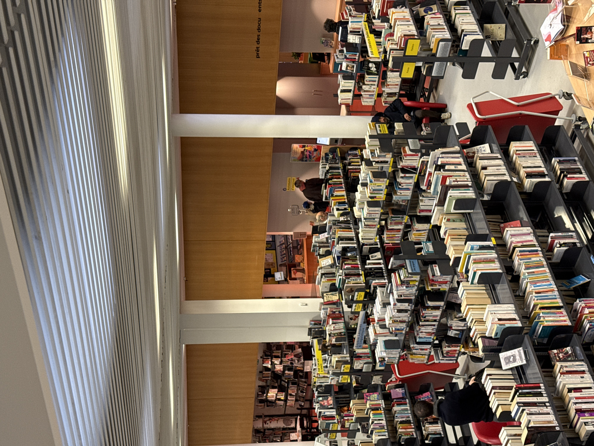 The image depicts the interior of a large, modern public library in Strasbourg, France. The scene is characterized by numerous rows of dark gray metal bookshelves filled with a wide variety of books, indicating different categories such as "700 Les arts & loisirs" (Arts & Leisure), "Espace Langues & Linguistique" (Languages & Linguistics Area), and "800 Littérature (Belles lettres)" (Literature - Fiction), denoted by yellow and black shelf markers.
The environment features a light-colored, possibly concrete floor and off-white structural columns. The ceiling is composed of horizontal, light-toned slats, with integrated lighting fixtures providing bright, even illumination. Higher sections of the walls are paneled with light brown wood.
At least seven individuals are present. In the lower-left foreground, a male subject in a dark top is seated in a red chair, facing away from the camera. In the mid-right, another male subject wearing a dark hooded jacket and light trousers is seated in a red chair, looking towards the left. In the mid-ground, near a sign reading "Automaties d'emprunt" (Borrowing Automats/Self-checkout), two individuals are visible, one in a dark jacket and another with a dark hat. In the far background, near an area marked "prêt des documents" (document lending), two subjects are standing by a counter, and another person is partially obscured by bookshelves in the far left background.
Observable objects include numerous books on shelves, red chairs, and a red folding utility cart with wheels in an aisle. Background areas show what appear to be computer terminals or kiosks for library services. People are engaged in activities typical of a library, such as browsing shelves or sitting, presumably reading or using devices. No direct interactions between subjects are clearly depicted.