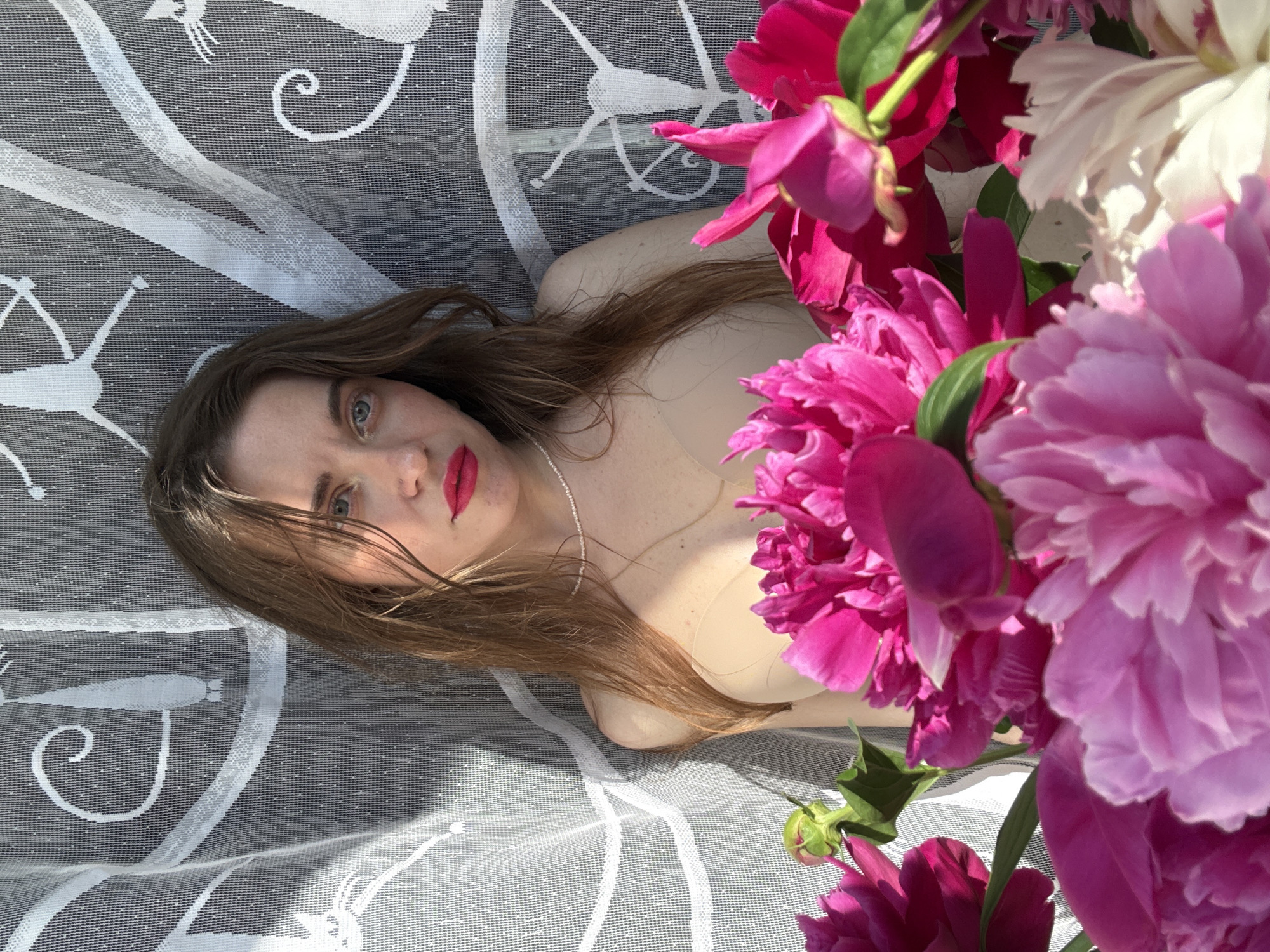 In an outdoor setting, possibly a balcony or garden, a young woman with long, light brown hair, wet from what appears to be recent showering, is positioned against a white lace curtain patterned with stylized cats. She is looking directly at the camera, her face accentuated with vibrant pink lipstick. She wears a nude-colored bra. In the foreground, a generous bouquet of peonies in various shades of pink and magenta obscures the lower half of the frame. The sunlight casts shadows through the lace curtain onto the woman and the background, creating a dappled effect. The location is identified as Unknown, Unknown.