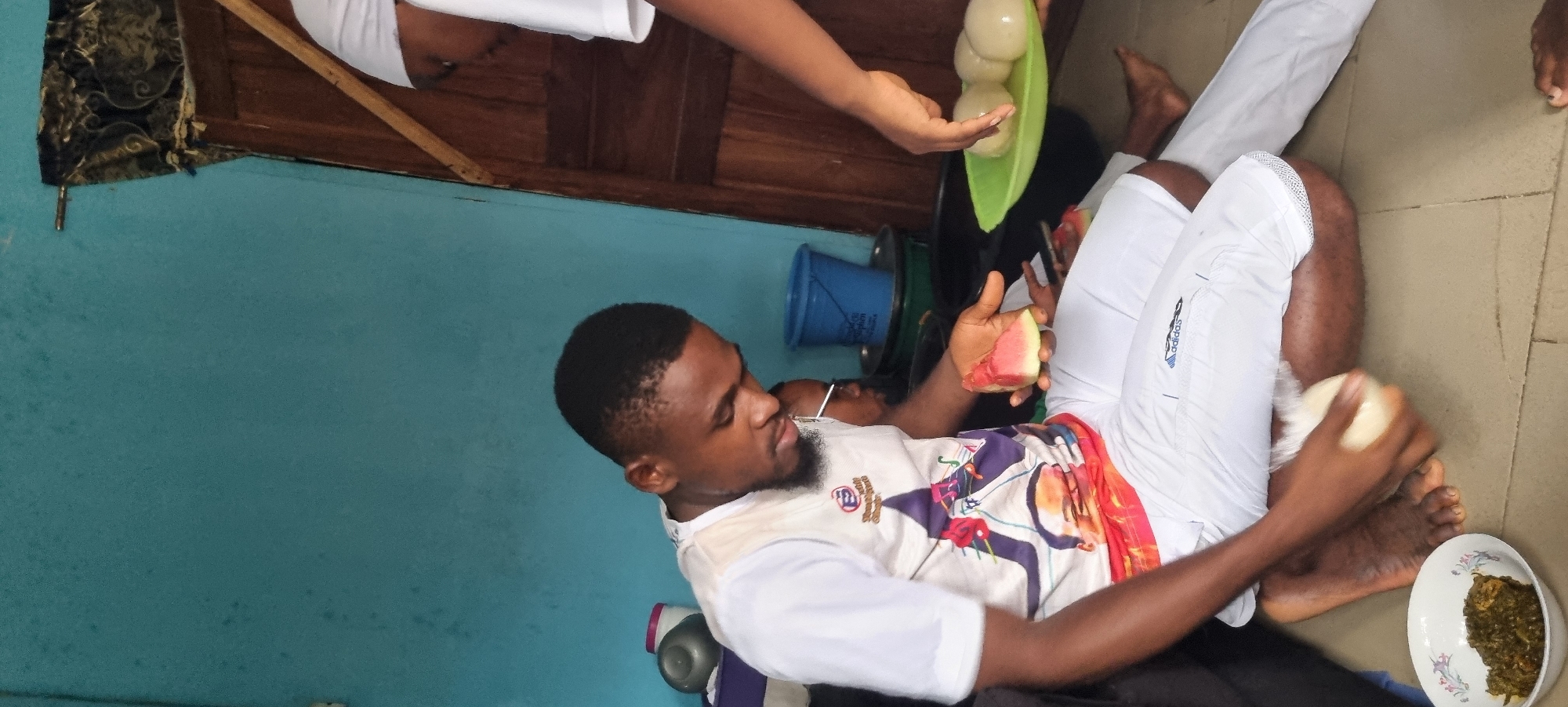 In Egan, Nigeria, a young man with dark skin and short hair, wearing a white t-shirt with a colorful graphic and white pants, sits on the floor. He is in the process of shaping a ball of white dough, likely fufu, with his hands. A slice of watermelon rests in his left hand. In front of him, a white bowl contains a green, leafy stew. To his right, another person, only partially visible, is offering a green plate filled with several balls of fufu. The background features a light blue wall, a wooden door, and various containers, including a blue bucket. The scene appears to be an informal gathering or meal preparation.