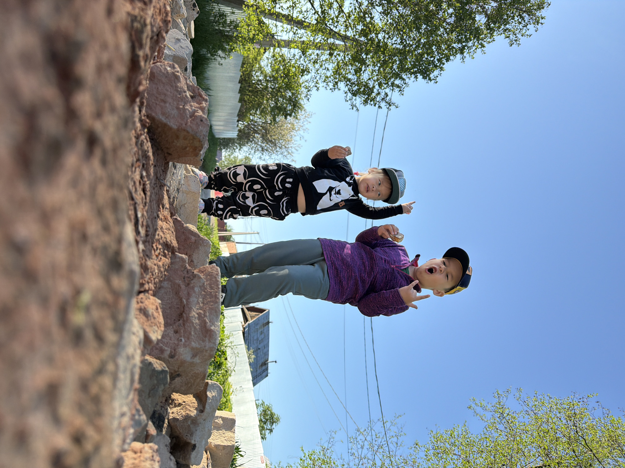 Two children pose on a rock wall. One child wears a black shirt with white graphics, black pants with white circles, and a dark baseball cap; they point. The second child wears a purple shirt, grey pants, and a dark baseball cap; they make a hand gesture and hold food. A rock wall is in the foreground. In the background is a blue sky, trees, a fence, a building, and power lines. Based on internet search results, it appears the image was taken in Chaikovsky, Russia.