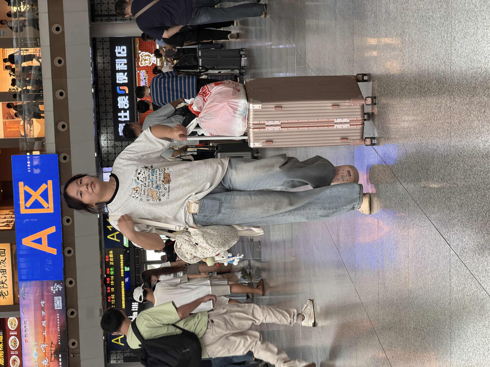 In Panyu District, China, a young woman is walking through a busy transportation hub, likely a train station or airport. She is moving from left to right in the frame, pulling a rose gold suitcase with her right hand. The suitcase has a plastic bag with red and white markings on top. She is wearing loose-fitting light blue jeans, a light-colored t-shirt with a cartoon print, and beige slide sandals with a heart pattern. On her left shoulder, she carries a small, light-colored bag adorned with a heart pattern and stuffed animals. Several other people are visible in the background, some with luggage, suggesting a travel environment. The floor is tiled and reflects the overhead lighting. Above, large digital screens display Chinese characters and advertisements, one prominently featuring the letter "A" and a stylized character. The overall scene depicts a moment of transit in a public space.