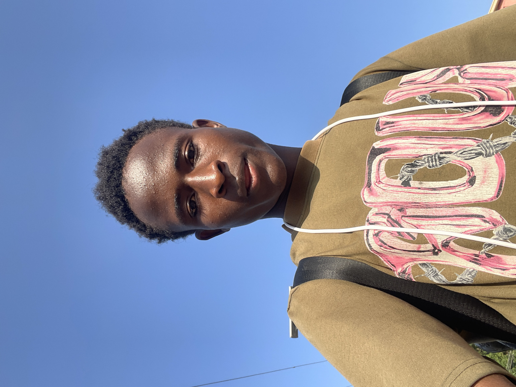 A young African male, with dark skin and short black hair, is pictured from the chest up. He is looking directly at the camera with a neutral expression. The sky behind him is a clear, bright blue. He is wearing a tan-colored t-shirt with a large, stylized graphic of pink lettering with barbed wire accents. A black backpack strap is visible over his right shoulder, and a white cord, possibly from headphones, hangs around his neck. The scene appears to be outdoors, possibly in a city environment, given the faint presence of a wire or pole in the upper left corner. There is no specific indication of the city of Jos, Nigeria, within the image itself.