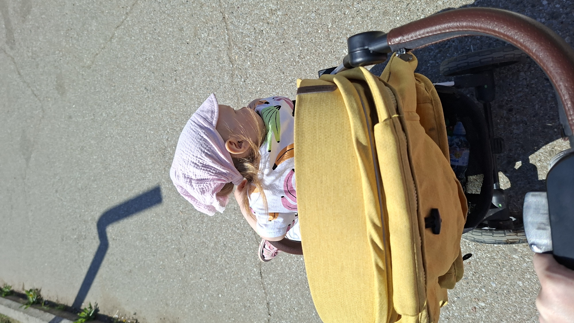 A high-angle shot shows a young child seated in a stroller on a paved surface, likely a sidewalk. The child wears a pink head covering, a t-shirt with a banana pattern, and light-colored shoes. The stroller has a brown handle and a yellow backpack hanging from it. The environment appears to be outdoors with asphalt or concrete visible. It is daytime, as evidenced by the bright lighting and shadows. The location is unspecified but identified as being in the city of Chaikovsky, Russia.