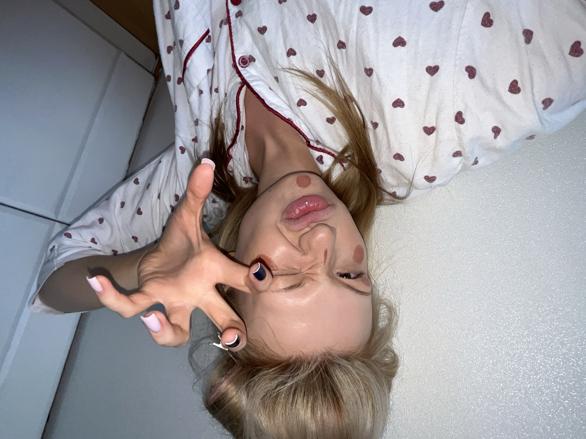 The image displays a close-up selfie of a young adult female, taken indoors within a residential setting in Chaikovsky, Russia. The subject has blonde hair, styled with volume on top and partially secured by a silver-colored hair clip visible near the crown. She is wearing a white pajama top featuring numerous small, dark red heart patterns and red piping along the collar and button placket. Her facial expression is exaggerated, characterized by a squinted left eye and puckered lips. Multiple reddish-brown circular marks are present on her left cheek, below her nose, and on her chin, with a faint reddish streak near her left eye. Her right hand is raised towards the camera, positioned in a claw-like gesture with fingers splayed and bent. The fingernails on her thumb, index, and middle fingers are painted light pink, while the ring and pinky fingers have dark blue/black polish with white tips. The background features a textured, light-colored wall on the left and a light gray or white paneled furniture piece, likely a wardrobe or cabinet, on the right.
