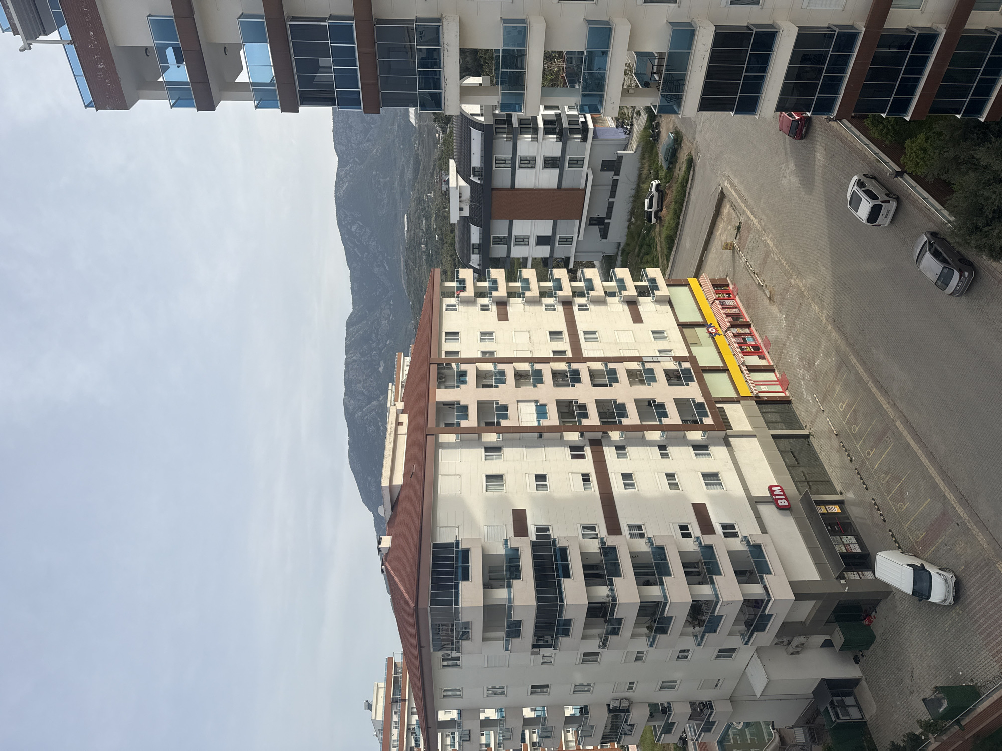 A slightly angled, aerial shot of a city block in Unknown, Unknown, features residential and commercial buildings against a backdrop of a mountain. The main building in the foreground is a high-rise apartment complex. Its exterior has a color scheme of white and brown with windows. The building includes a BİM storefront at street level, recognizable by its red logo. A white van is parked in front of the BİM store.
Other buildings are visible beyond the main high-rise, including another modern apartment building on the left edge. Between buildings, there are some greenery and additional residential buildings.
A parking lot and street are adjacent to the high-rise. Several cars of different colors are parked or moving on the street.
In the background, a large mountain occupies the central-left side. The sky is light with visible clouds.