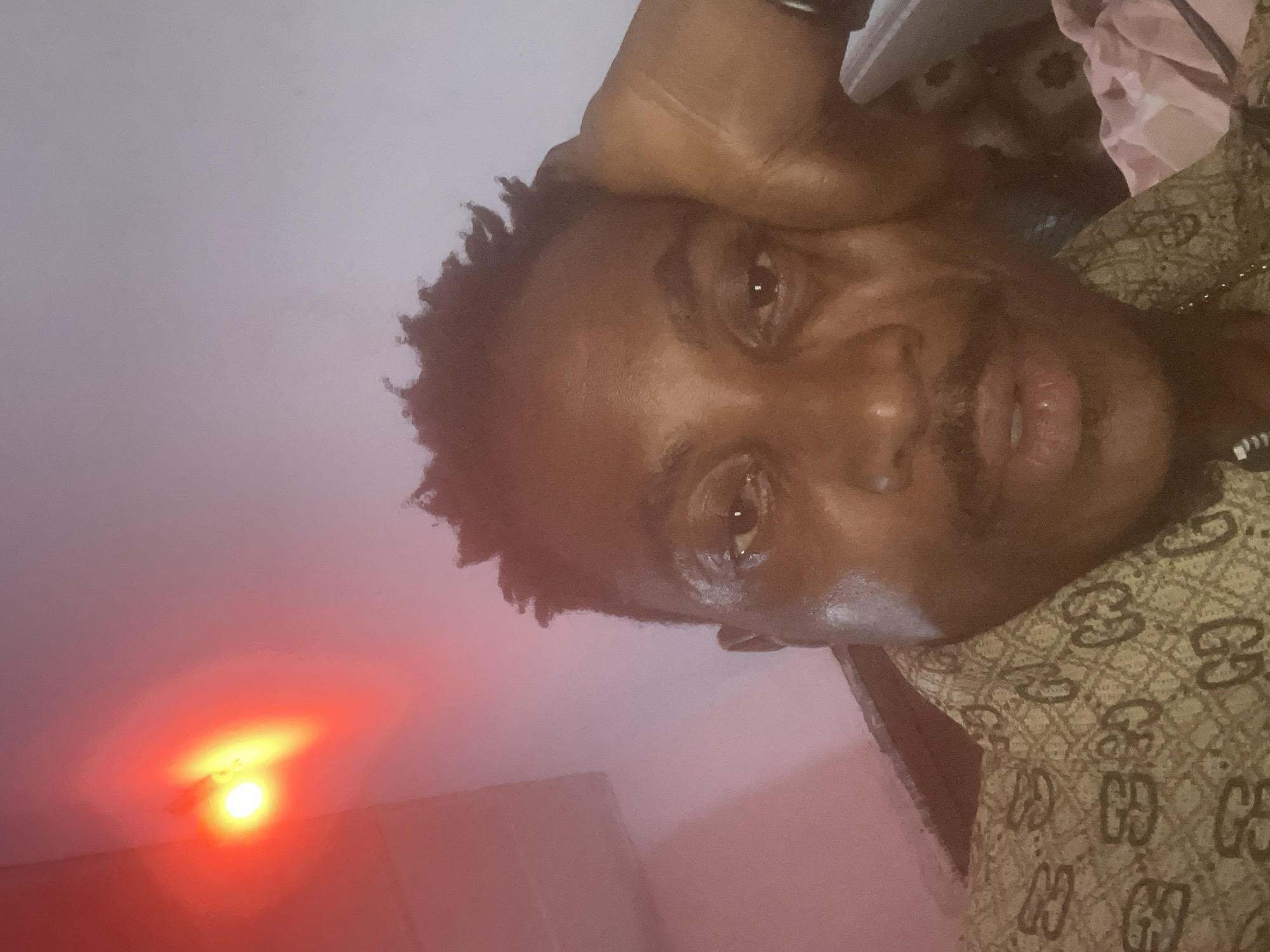 The image is a close-up selfie of a Black man in Adankolo, Nigeria. He is wearing a light brown shirt with a repeating pattern of dark brown interlocking "G" shapes. His right hand is resting on his cheek, and his left eye is visible, looking towards the camera. His lips are slightly parted, and he has a mustache. In the upper left corner of the image, there is a bright red light source with a yellow glow. The background appears to be a plain wall with a reddish tint in some areas, possibly from the red light. The overall lighting suggests an indoor setting, possibly at night or in a dimly lit room.
