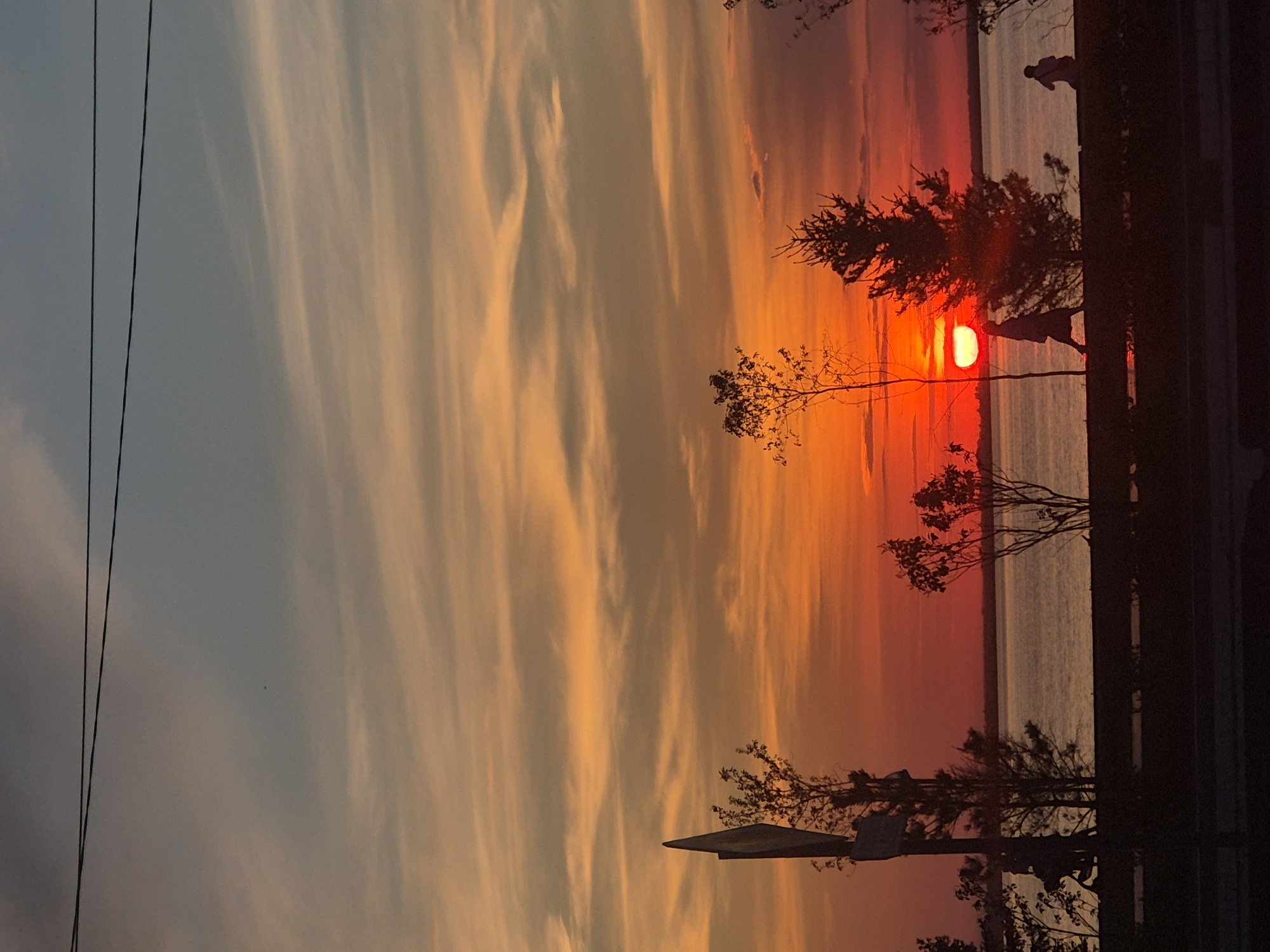 This is a photograph of a sunset over a body of water, likely a lake or a bay, in Kokshetau, Kazakhstan. The sky is filled with vibrant colors, ranging from deep orange and red near the horizon to softer hues of yellow and blue higher up. Thin, wispy clouds stretch across the sky, catching the light of the setting sun. Silhouetted against the bright sky are trees and a street lamp on the left side of the frame. In the middle ground, a person is walking along a path next to the water. Further in the distance, another person is seated. The water is calm, reflecting the colors of the sunset. The overall scene evokes a peaceful and serene atmosphere.