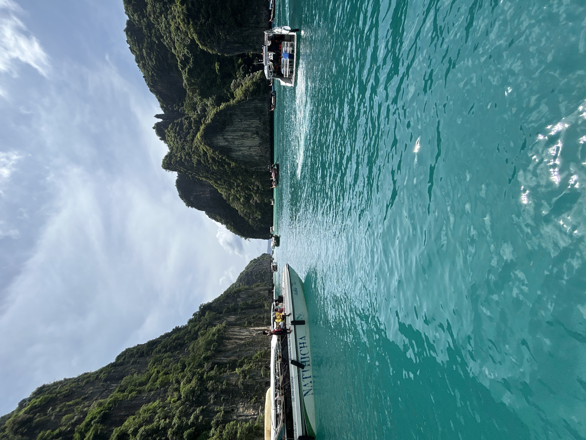 This is a vertical, eye-level, wide shot of Ao Nang, Thailand, at midday. The foreground is dominated by turquoise water, with ripples and reflections from the sun. In the middle ground, several longtail boats and speedboats are visible, some with passengers. One speed boat, named "NATTHICHA," is prominently featured on the left, with several individuals on board. The background consists of towering, lush green limestone cliffs that frame the scene. The sky above is a mix of blue and white clouds, indicating fair weather. The overall impression is a busy, tropical tourist destination.