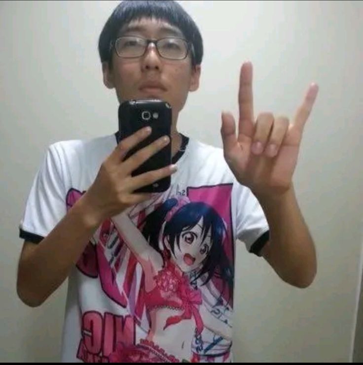 A young man with short black hair is standing in front of a mirror, wearing a white t-shirt with a black trim and a large image of a female anime character printed on it. He is holding a black smartphone in his left hand, and his right hand is raised in a rock and roll hand gesture. He is facing the mirror and looking at his reflection. The image appears to be taken in a bathroom, as the background is a plain white wall, and there is a light fixture in the upper right corner. The image is taken from an angle slightly above the man's head. The location of the image is a residential area in the city of Geneng, Indonesia, based on coordinates (latitude: -7.45472, longitude: 111.427584).