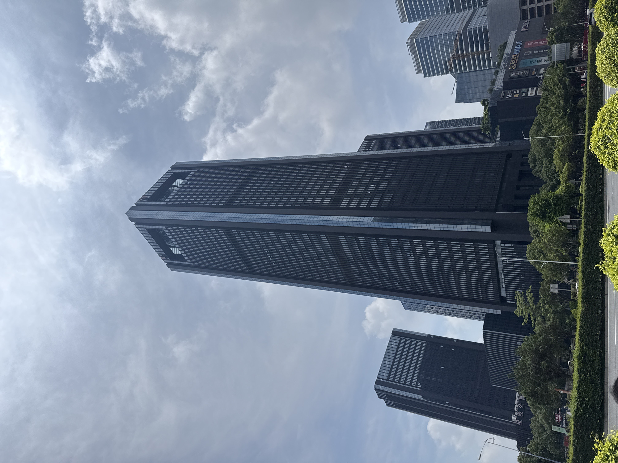 This image depicts a modern urban landscape in Haizhu District, China, under a bright, partly cloudy sky. The central feature is a prominent, dark-paneled skyscraper with a distinctive open-frame structure at its apex, featuring vertical light-colored accents. To the left, another large, dark, glass-clad building appears at an angle, while several other multi-story commercial and residential buildings with varied architectural styles are visible in the background on the right. Below these structures, a low-rise commercial complex is evident, displaying various retail signs and logos, including "VMAX", "McDonald's", and a large "IMAX" sign. The foreground features a multi-lane road bordered by well-maintained, dense green shrubbery and mature trees. Several streetlights are positioned along the green median. The natural lighting suggests daytime conditions. No specific individuals or active interactions are discernible within the frame.