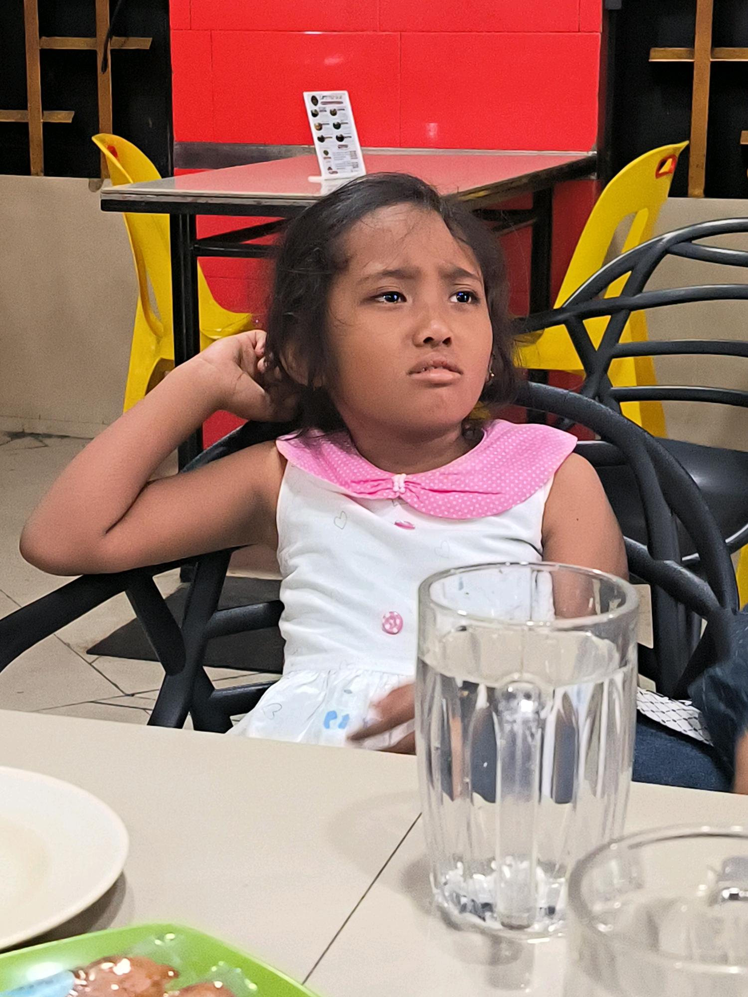 A young girl with dark hair, wearing a white dress with a pink collar, sits at a table in a restaurant. Her right arm is propped up, and her hand is near her ear, as she looks away from the camera with a thoughtful expression. In front of her is a glass of water and partially visible plates of food. The background features red walls, yellow chairs, and black table structures, suggestive of a casual dining establishment. The flooring is tiled with a geometric pattern. While the precise location within Banda Aceh, Indonesia, is not discernible from the visual cues, the restaurant setting is evident.