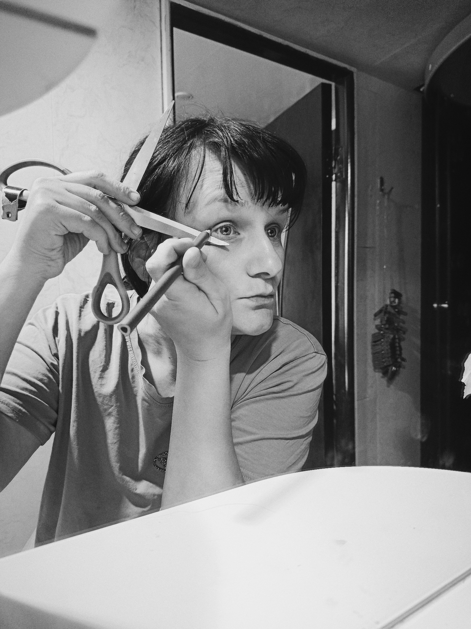 A black and white photograph depicts a person engaged in self-grooming activities, seemingly in an indoor setting like a bathroom or vanity. The central subject, a person with short, dark hair, is positioned in front of a mirror, with their focused gaze directed at their reflection.
The person is actively using two implements: a pair of scissors held in their left hand, with the blades open and positioned near their forehead, suggesting they are cutting or trimming their own hair or bangs. The scissors have a distinctive ergonomic handle, with one loop being an elongated, almost teardrop shape. Simultaneously, their right hand holds a dark, pencil-like object close to their right eye, indicating they are applying makeup, likely eyeliner or kohl.
The subject wears a light-colored, short-sleeved t-shirt, on which a small, possibly embroidered, detail or emblem is visible on the chest. A white counter or table surface occupies the lower right foreground of the image.
In the background, a doorway is partially visible on the right, leading into a darker space. On the wall beside the doorway, a small, dark, and intricate decorative item or hanging object is discernible. The overall lighting is soft but creates strong contrasts typical of black and white photography, highlighting the textures of hair, skin, and fabric. The expression on the person's face is one of intense concentration. There is no visible text in the image. The specific location and time of day are unknown, though it is clearly an indoor environment.