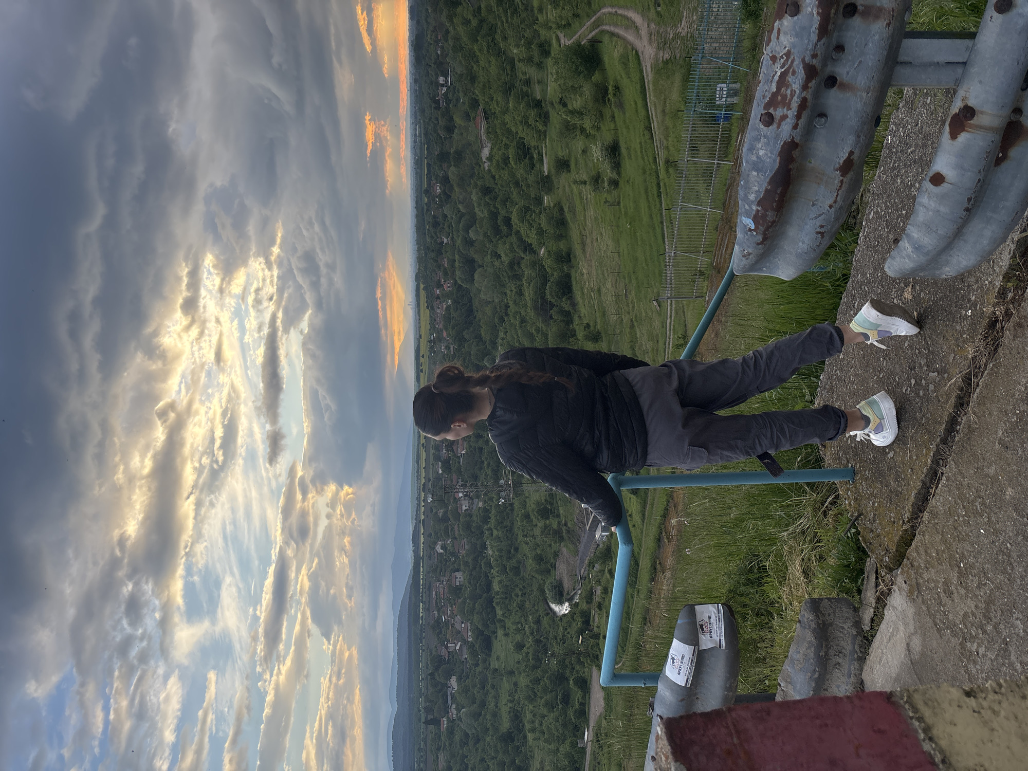 A woman with a long ponytail, dressed in a dark jacket and gray pants, stands on a concrete structure with rusty metal railings, overlooking a vast landscape in Ognyanovo, Bulgaria. The view encompasses a green valley with a town nestled within it, and mountains in the distance. The sky is filled with dramatic clouds, lit by the setting sun. She wears white sneakers with light green and purple accents. The image captures the elevated perspective and the scale of the surrounding environment.
