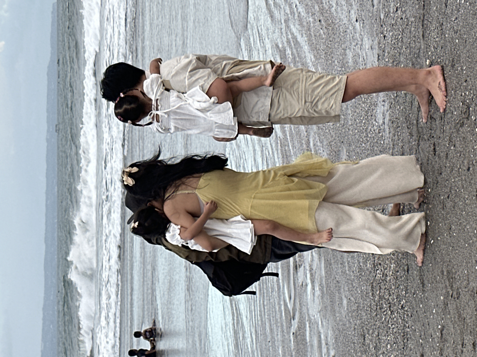 Two couples are standing on a beach in North Kuta, Indonesia, facing the ocean. The couple on the left consists of a woman in a yellow dress and a man in a dark jacket holding a young girl in a white dress. The couple on the right consists of a man in a light-colored shirt and beige shorts, holding a young girl in a white dress. The ocean has large waves crashing. In the background, two young children are sitting in the shallow water. The beach is wet sand with small shells. The sky is overcast.