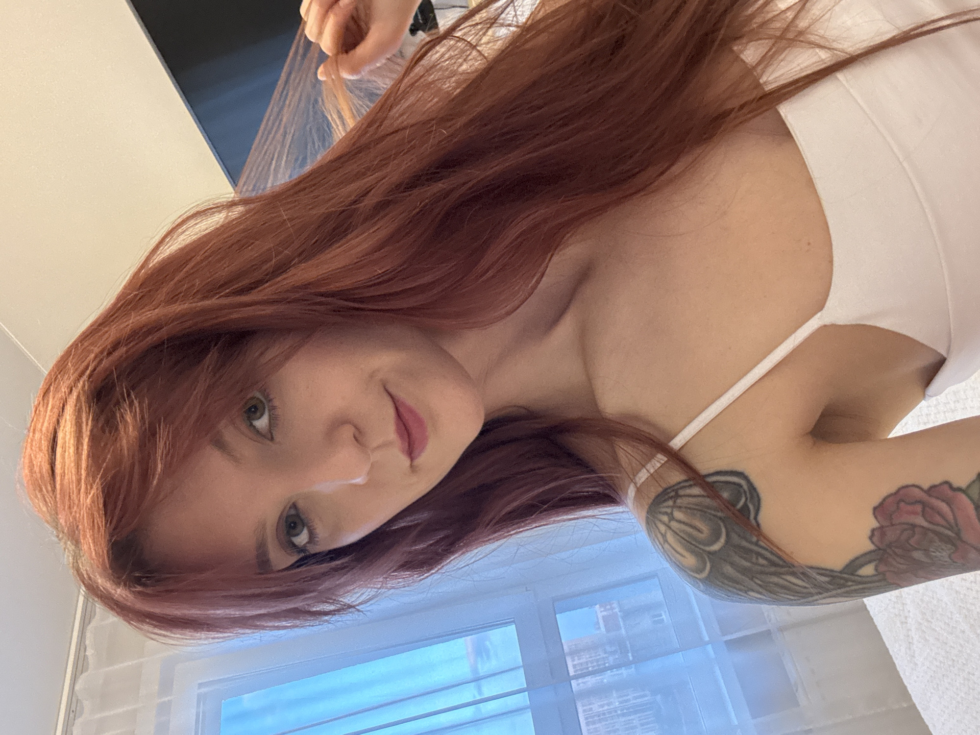 The image features a woman with auburn hair and light skin. She is wearing a white tank top. A visible tattoo adorns her left arm. The background shows a window with white blinds, revealing a cityscape. Based on the urban architecture visible through the window, the location may be Izhevsk, Russia, but that cannot be confirmed with certainty. There is no interaction or action occurring. The woman is simply looking at the camera. A hand, possibly her own or someone else's, can be seen near her hair.