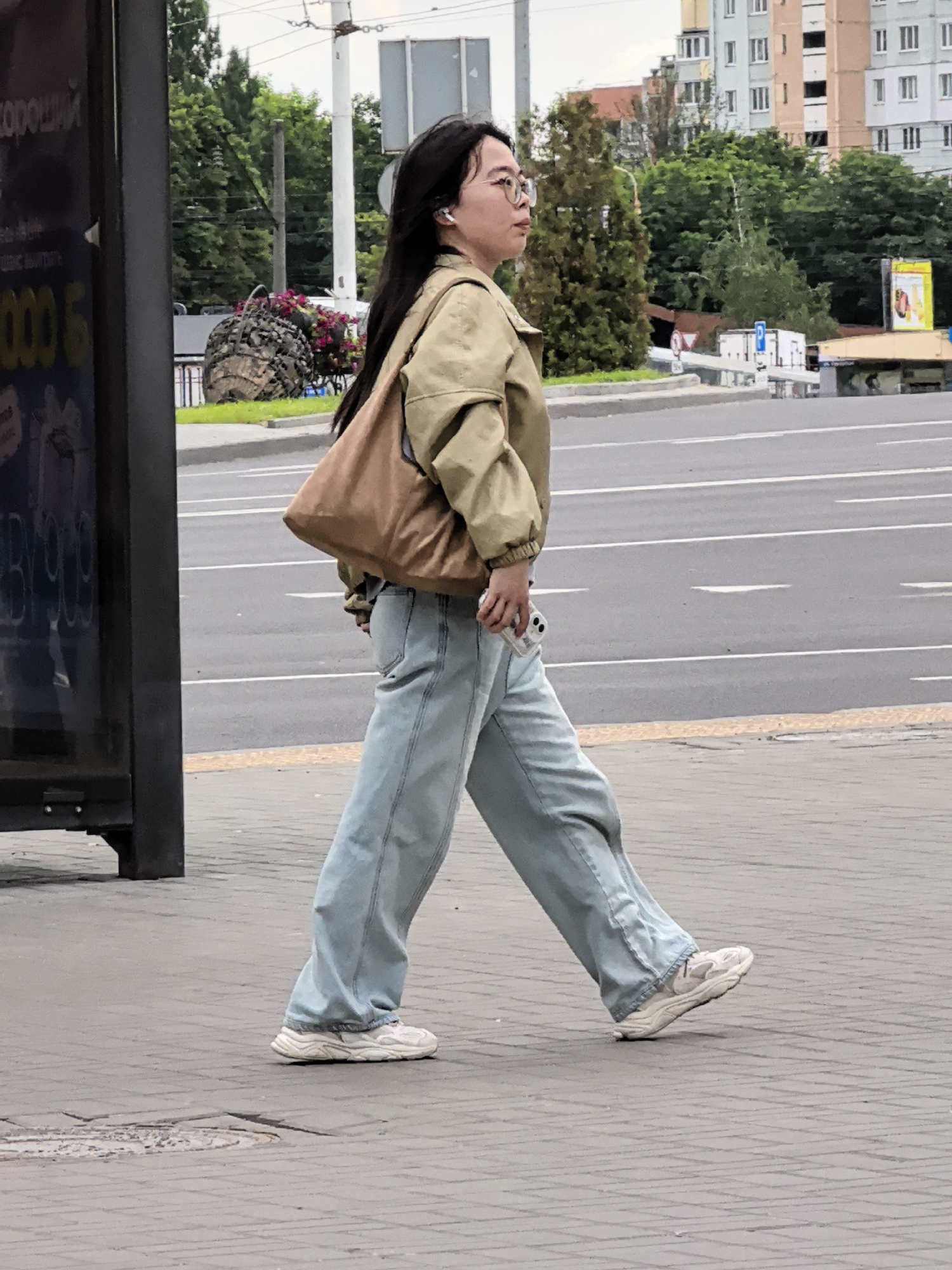 A person with dark hair and glasses walks on a sidewalk next to a street in an urban area. The person is wearing a tan jacket, light blue wide-leg jeans, and white sneakers. They are carrying a brown shoulder bag and holding a white smartphone in their right hand. In the background, there are trees, buildings, a bus stop shelter, and traffic lights. The weather appears overcast.