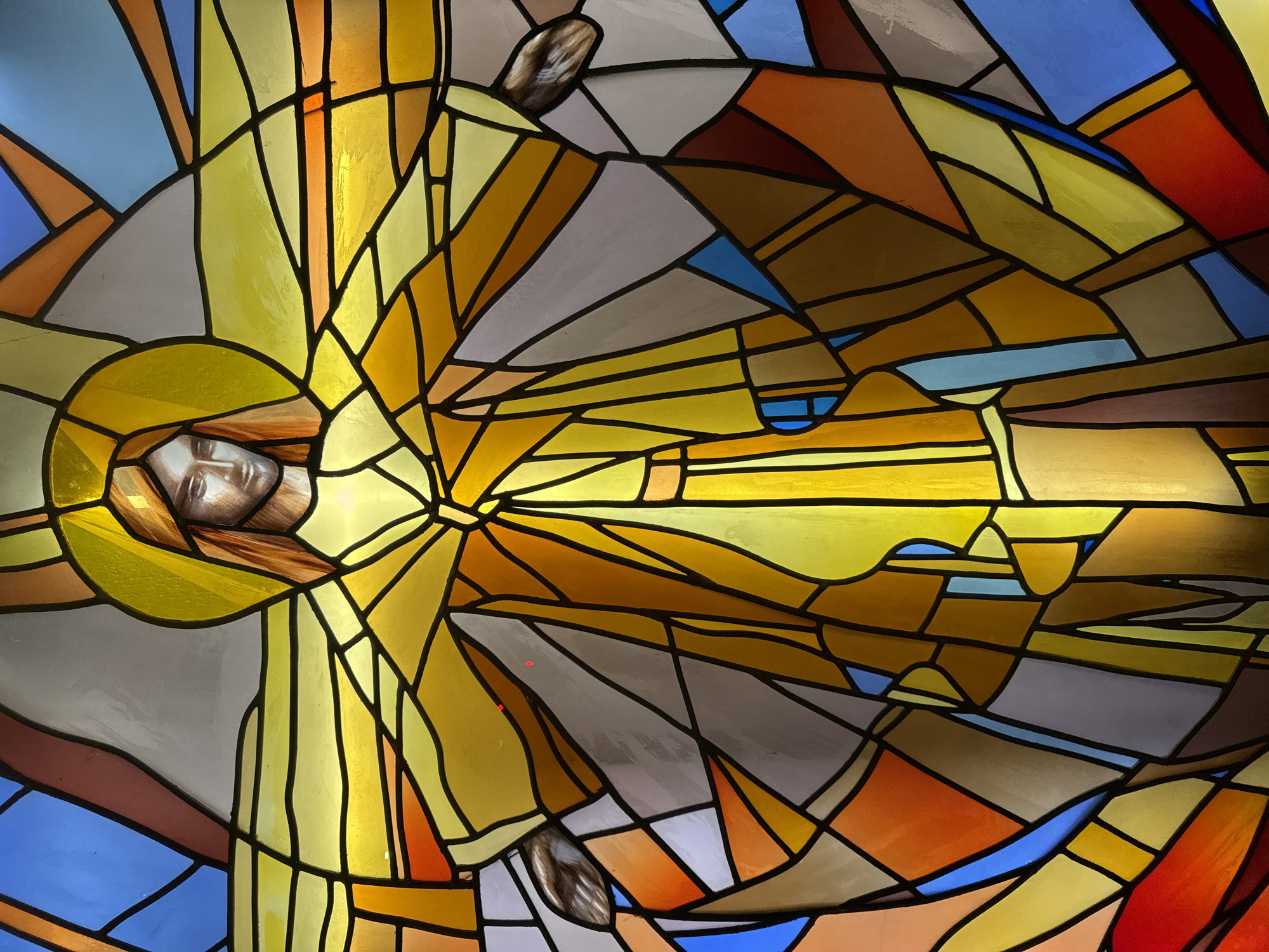 This image is a close-up of a stained-glass window depicting a figure, likely a religious figure, emanating light. The face of the figure is realistically rendered and surrounded by a golden halo. The rest of the figure and the background are abstracted into geometric shapes filled with varying shades of yellow, gold, grey, orange, blue, and red. The design suggests light radiating outward from the central figure. There is no specific location information provided; the information about Paris is not confirmable.