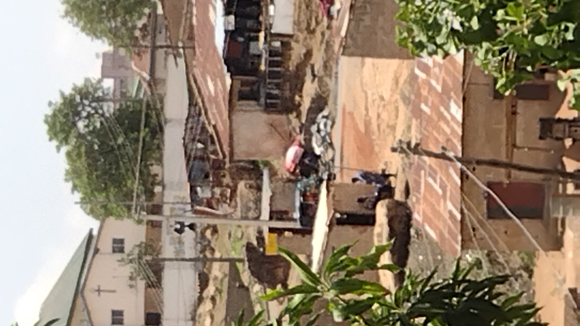 The image shows an urban scene in Jos, Nigeria, viewed from an elevated perspective. The scene features buildings with a mix of corrugated metal roofing and painted walls, interspersed with foliage. There is evidence of disrepair and informal settlements. Power lines are visible. A church is visible in the background. Personal items and debris are scattered throughout the scene.