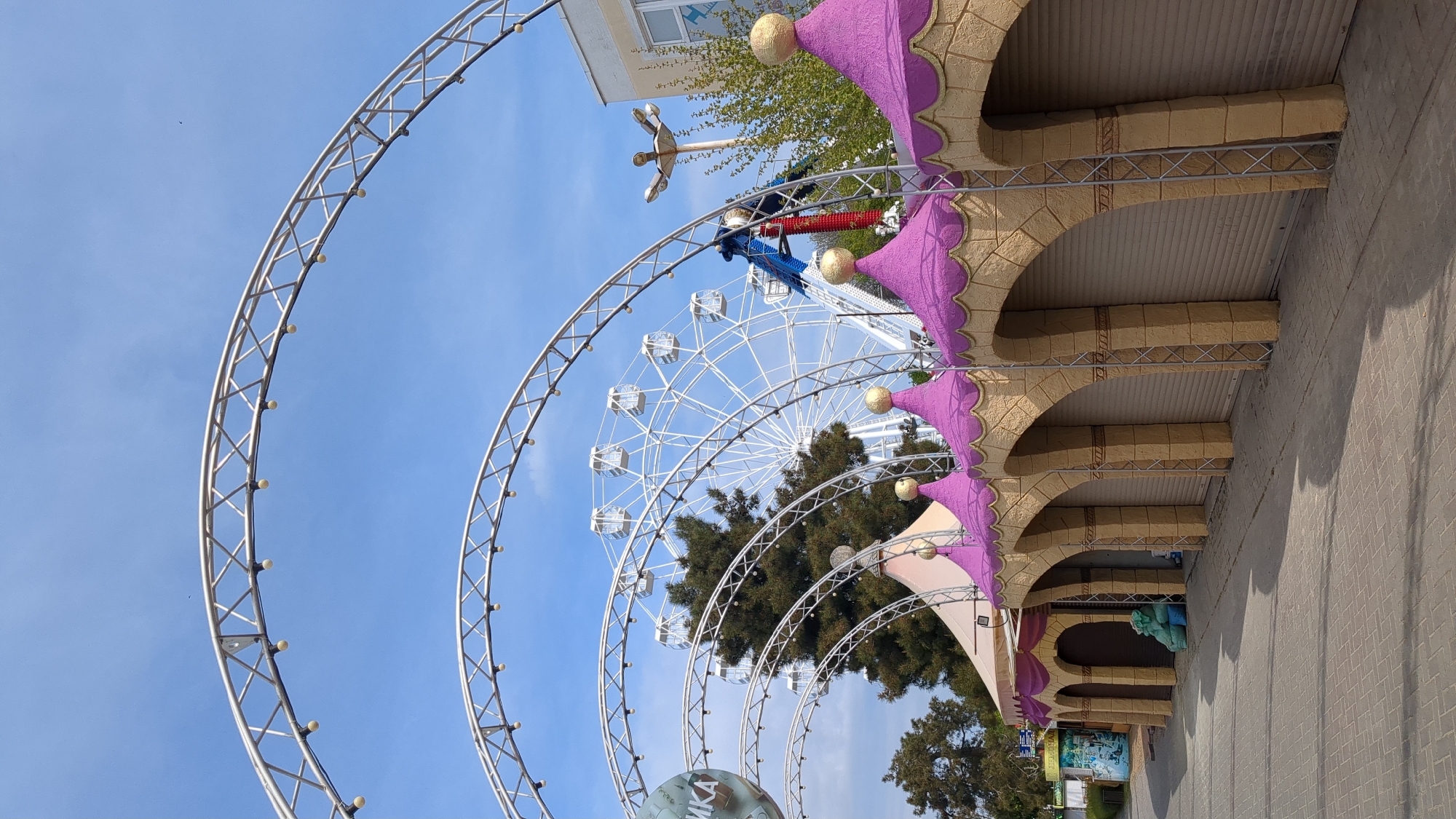 This vertical shot captures a vibrant amusement park scene under a clear blue sky, likely in Gelendzhik, Russia, as suggested by the context. The dominant feature is a large, white Ferris wheel positioned towards the center-right of the frame, its many cabins clearly visible.
In the foreground, a series of gleaming silver metal archways, resembling truss structures with embedded lights, curve from the left side of the image towards the center and right. These archways appear to form an entrance or a decorative tunnel. To the right, a row of elaborate buildings or stalls with distinct architectural features is visible. These structures feature light-brown, stone-like arches and ornate purple, scalloped roofs topped with decorative golden spheres.
Beyond the foreground archways and to the left of the Ferris wheel, another tall amusement park ride with a red and blue mechanism and a propeller-like top can be seen. Lush green trees are interspersed throughout the park, adding natural elements to the colorful attractions. The ground is paved with light-colored, cobblestone-like bricks.
The time of day is clearly daytime, characterized by bright, even lighting and a largely cloudless blue sky, with only a few wispy clouds. A small dark bird is visible flying in the sky in the upper left section. On a large sphere-like object partially visible on the ground beneath the metal arches, the Russian word "АРКА" (ARKA, meaning "arch") is visible. Other partially obscured Russian text can be seen on signs beneath the arched buildings, including "КАТАЛОГ" (KATALOG). The overall impression is one of an inviting and playful environment, typical of an amusement park.