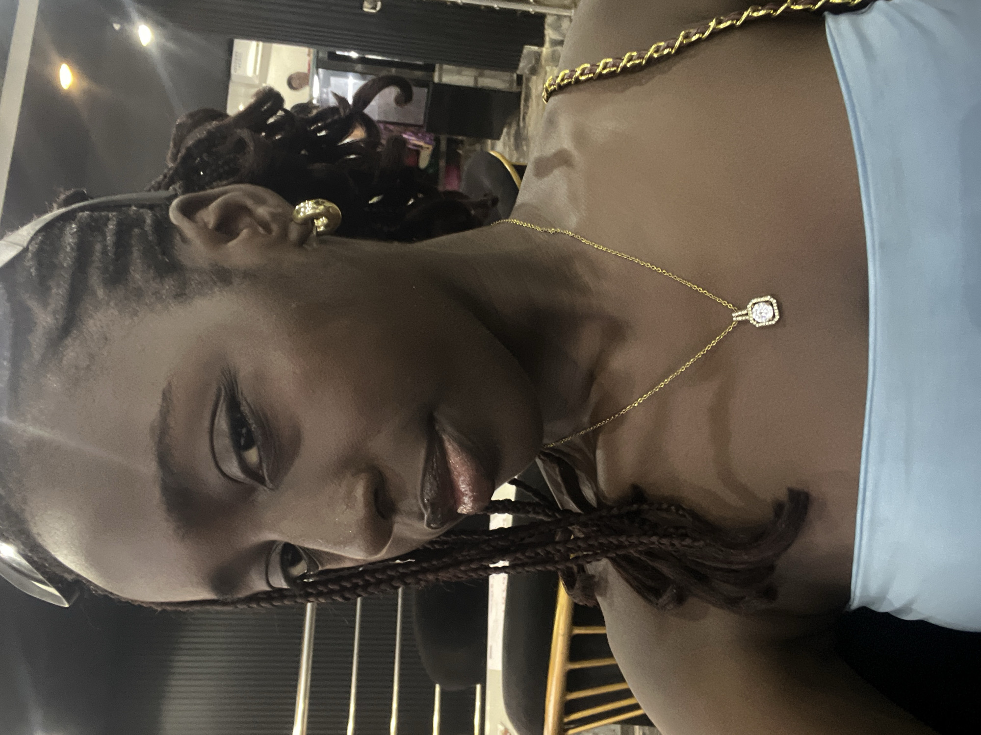 A young Black individual, likely female, is captured in a medium close-up, gazing directly at the viewer with a slight smile. She has dark braided hair, with sunglasses resting on top of her head. Her eyes are accentuated with dark eyeliner, and her lips have a glossy, reddish-brown tint. She wears a light blue strapless top. Prominent accessories include a chunky gold hoop earring on her right ear and a delicate gold chain necklace featuring a square pendant with a large clear central stone surrounded by smaller clear stones. The shoulder strap of a bag, composed of a gold chain interwoven with dark material, is visible over her right shoulder. The background indicates an indoor setting, possibly a commercial establishment in Abuja, Nigeria, characterized by a dark, vertically textured wall on the left, partial views of wooden chairs, and reflected overhead lighting. A blurred human figure is faintly visible in the far-right background.