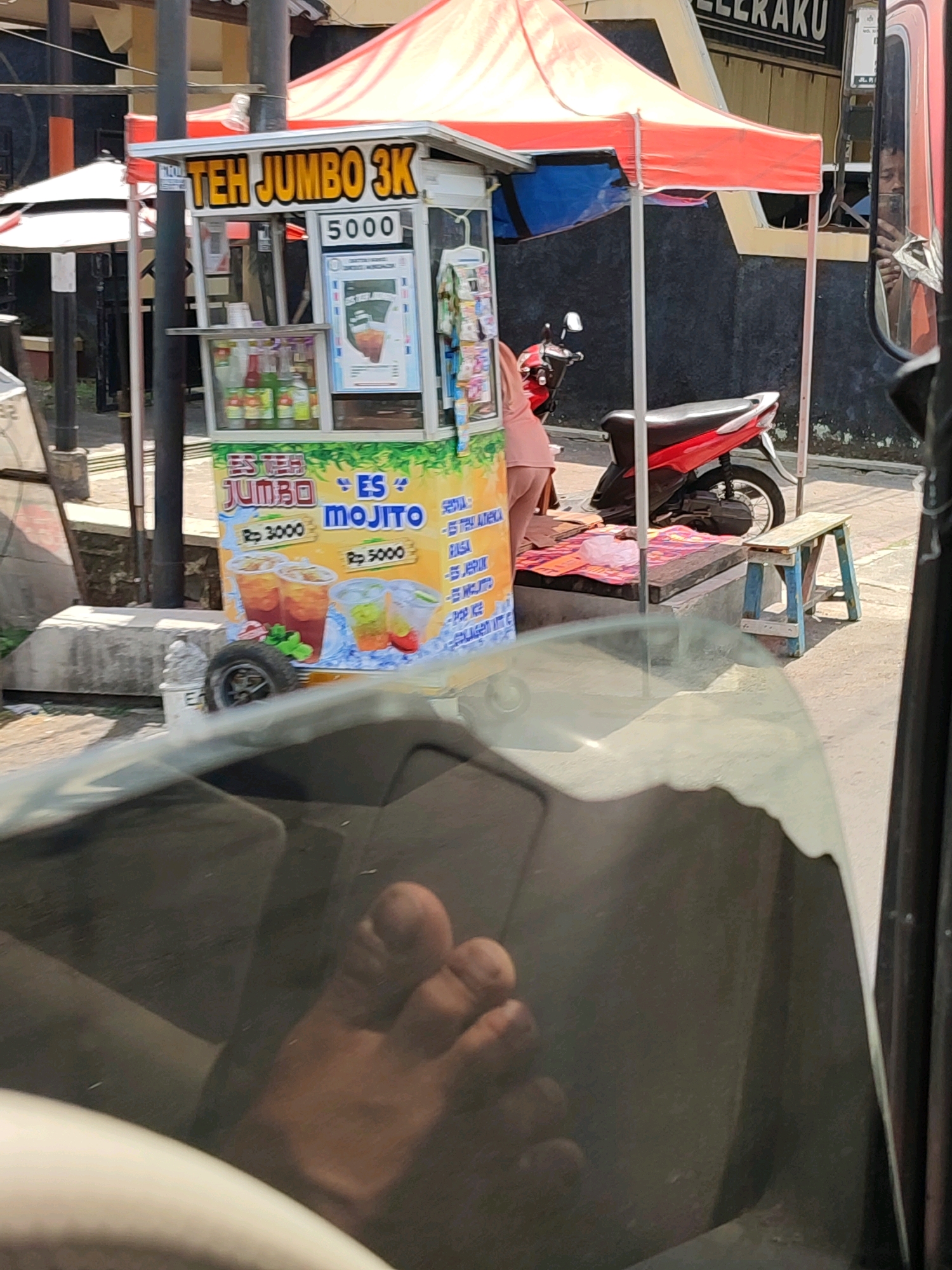 The image depicts a street scene in Magelang, Indonesia, featuring a beverage cart selling "Teh Jumbo 3K" and "Es Mojito". The cart is adorned with colorful advertisements showcasing drinks and prices, with "Rp 3000" and "Rp 5000" visible. A red motorcycle is parked behind the cart, and a small blue and white stool is placed nearby. In the foreground, a bare foot is visible, suggesting the perspective of someone inside a vehicle. The environment appears to be a street with paved ground and a building in the background. The overall scene captures a typical street vendor setup in an Indonesian urban setting.