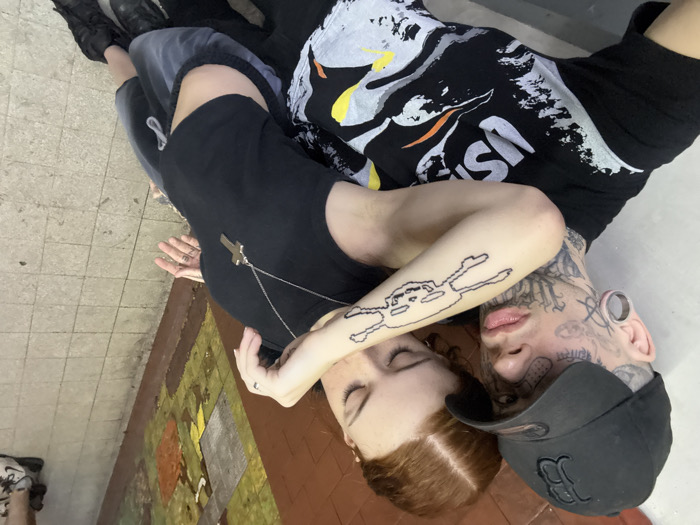 Two people lie on a tiled floor. One person with red hair wears a black shirt with a cross necklace and has a tattoo on their arm. The other person has extensive face and neck tattoos, ear gauges, and wears a black baseball cap. Both are lying down, close to each other. Additional people's legs and feet appear in the background.