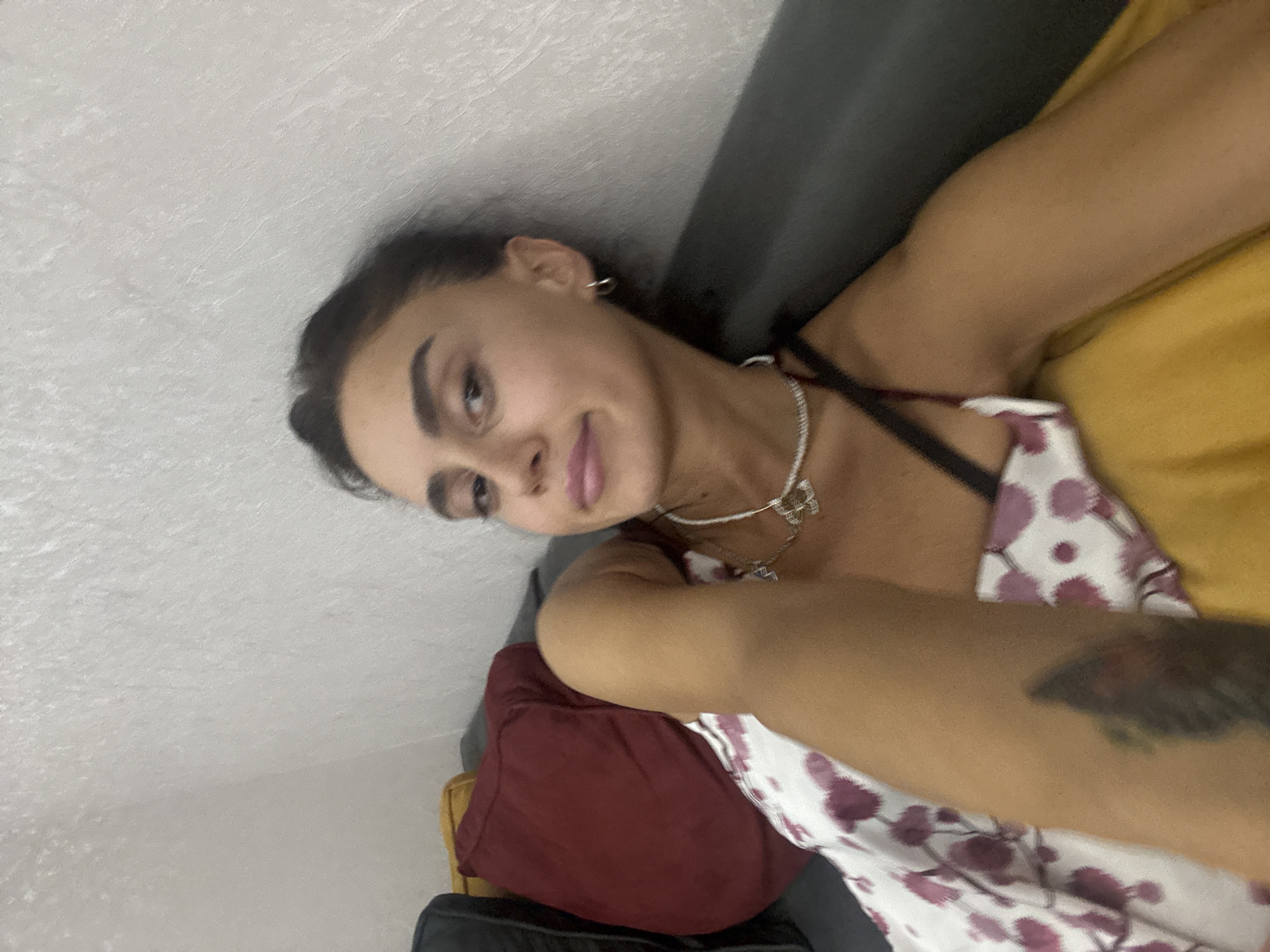 A young woman with dark hair is lying on a sofa, leaning against a textured, off-white wall. She is wearing a white dress with a pink floral pattern and a black bra strap is visible. She has a delicate silver necklace with a pendant around her neck and small hoop earrings. Her left arm, which is resting on her chest, has a tattoo. The sofa is dark gray, and there are two pillows behind her: one is deep red and the other is mustard yellow. The scene appears to be indoors, likely in an apartment in Tbilisi, Georgia.