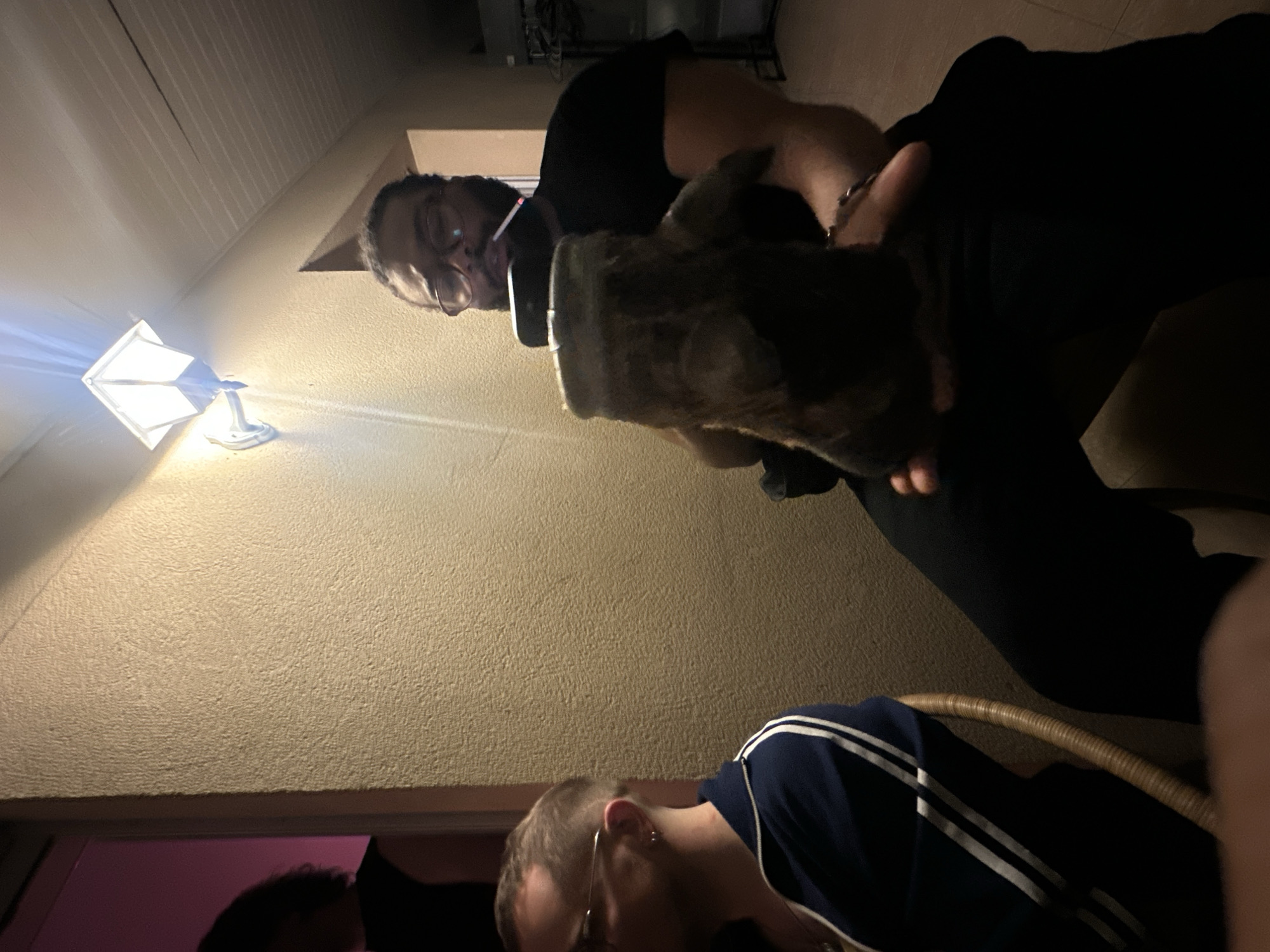 The image shows an indoor scene with two people and what appears to be a small animal. The man in the foreground has blond hair and wears a blue shirt with white stripes. The other man in the background wears glasses, a black shirt, and dark pants. He is holding a furry animal. The image is taken from a low angle, looking up. On the wall behind them is a light fixture. The background suggests an indoor or enclosed outdoor space with a textured wall. The location is Montreuil, France.
