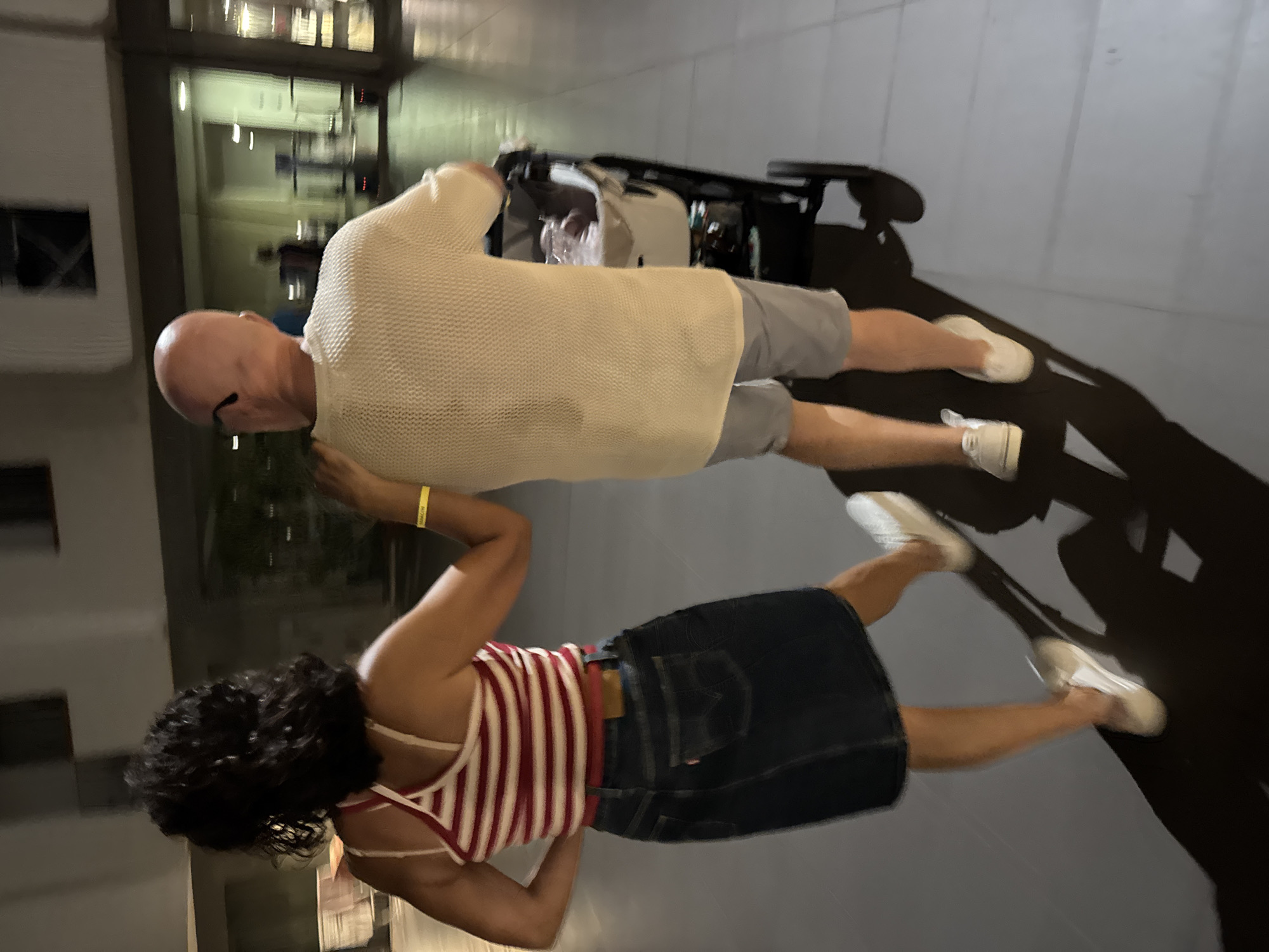 In Isola delle Femmine, Italy, two individuals are walking with a stroller at night. A woman with dark, curly hair, wearing a red and white striped tank top and a denim skirt, walks slightly ahead and to the left, her left arm resting on the shoulder of the man beside her. The man, bald with glasses, is dressed in a light-colored, textured sweater and grey shorts. He is pushing the stroller, which contains a baby. The background shows a blurred, modern building with illuminated windows and doorways. The pavement is tiled.