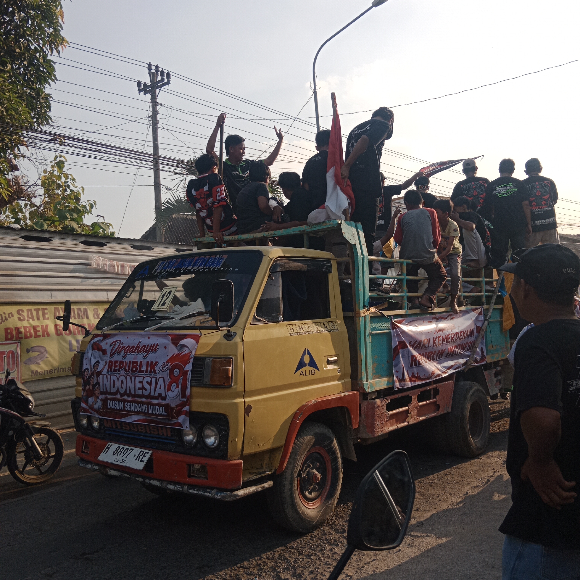 The image captures a daytime scene in Tegalsari, Indonesia, likely during a parade or celebration. A yellow Mitsubishi truck is the central focus, adorned with banners that read "Dirgahayu Republik Indonesia 81." The truck is filled with a group of young men, many wearing black t-shirts, some with logos. Some are standing, others are seated on the truck's edge. One individual is waving a red and white Indonesian flag.
In the background, electrical poles and wires are visible, along with trees and structures that suggest an urban or semi-urban environment. A motorcycle is parked on the left side of the frame. Another banner, this one yellow, is partially visible on the left, advertising "Sate Ayam & Bebek Goreng". The overall atmosphere suggests a festive or patriotic event.