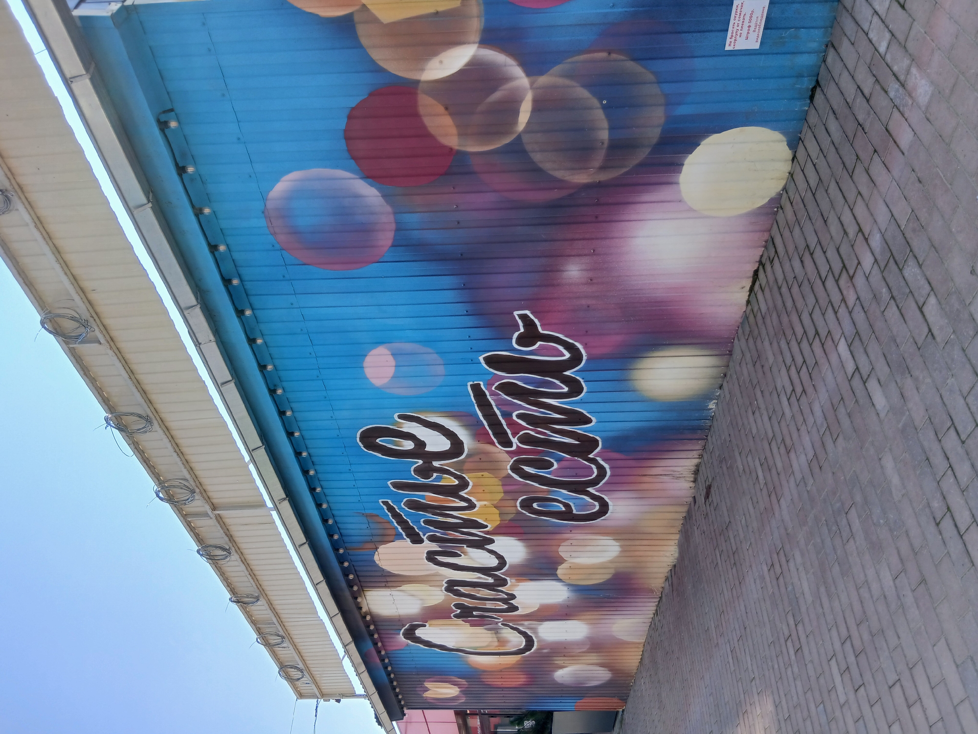 A mural is painted on a blue corrugated metal wall in the city of Osa, Russia. The mural features large, stylized white text with dark outlines on a background of colorful, blurred circles resembling bokeh lights. The text appears to be in Russian, with the prominent words "Счастье есть" (Happiness exists). The wall is part of a structure that extends horizontally, and there are lights mounted above the mural. To the right of the mural, a small white sign with Russian text is attached to the wall. In front of the wall is a paved walkway made of reddish-brown rectangular bricks. The sky above is clear and blue. The scene depicts an outdoor urban environment.
