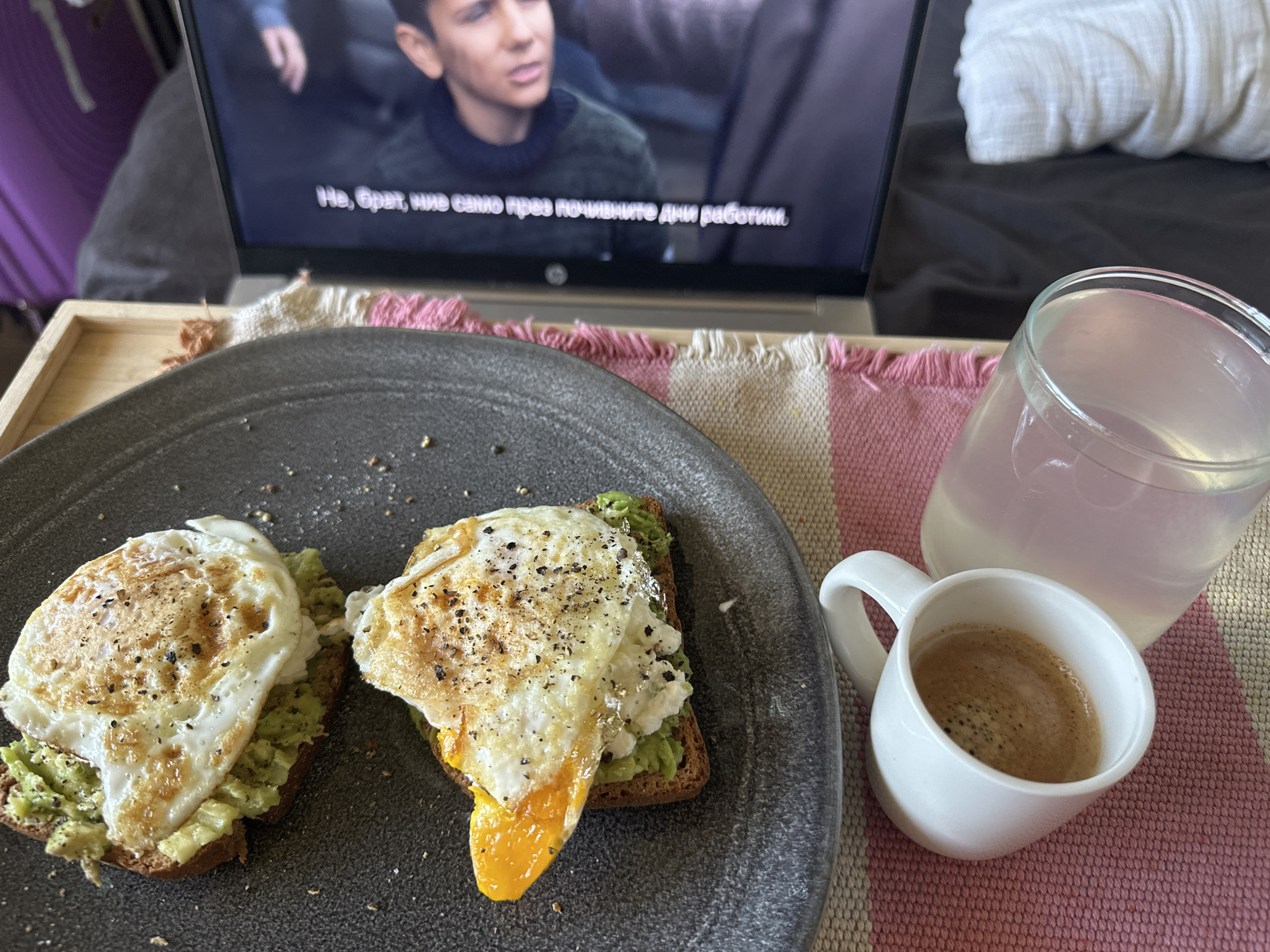 A close-up shot of a meal setting. Two slices of toast topped with mashed avocado and fried eggs are placed on a gray plate. A small white mug filled with coffee and a glass of liquid stand beside the plate. In the background, a laptop displays a movie with subtitles in Bulgarian. The scene is set on a striped pink and white mat, possibly on a bed tray. No people are visible in the shot. No indications point to the city of Sofia, Bulgaria.