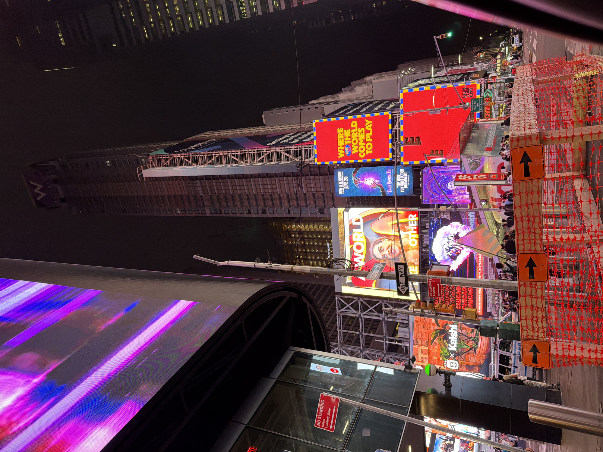 The image depicts a nighttime street scene in Times Square, New York City. Prominent features include a large, illuminated billboard displaying a pink and purple abstract design on the left, and a tall skyscraper with a "W" logo at its peak in the background. Several other billboards with various advertisements are visible, including one with the text "WHERE THE WORLD COMES TO PLAY" in bright red. A street light with a green bulb is illuminated. A police van is parked on the street, and a crowd of people are walking along the sidewalk. Orange construction netting with directional arrows is set up in the foreground, indicating ongoing work. The overall atmosphere is one of urban vibrancy and commercial activity, characteristic of Times Square at night.
