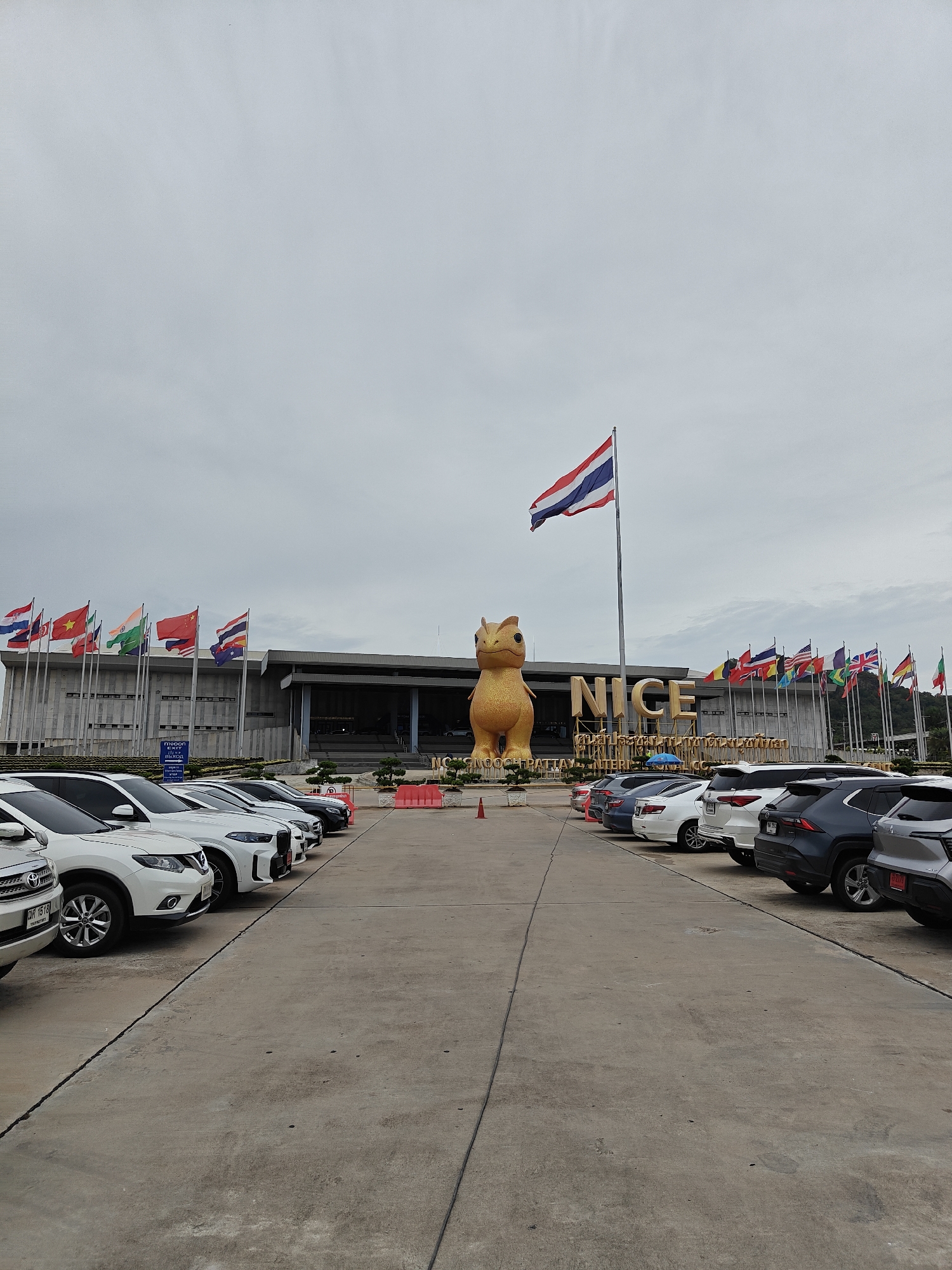 This is a low-angle, eye-level, wide shot of the outdoor area in front of the NICE (Nong Nooch International Convention and Exhibition) Center in Na Chom Thian, Thailand. The sky is overcast. In the center of the image, a large, golden, stylized dinosaur statue stands prominently. Behind the statue, the NICE center building is visible, with its name displayed in large, gold lettering. A Thai flag flies high on a flagpole in front of the building, to the left of the statue. To the left and right of the building, numerous national flags from various countries are displayed on poles, suggesting an international gathering or presence. In the foreground, a parking lot is filled with a row of cars, predominantly white SUVs and sedans, parked in organized lanes. The ground is paved with concrete, and there are some orange traffic cones and red barriers placed in the parking area. The scene appears to be a public space, likely a tourist attraction or convention venue, with a clear emphasis on international representation. No people are visible in the image.