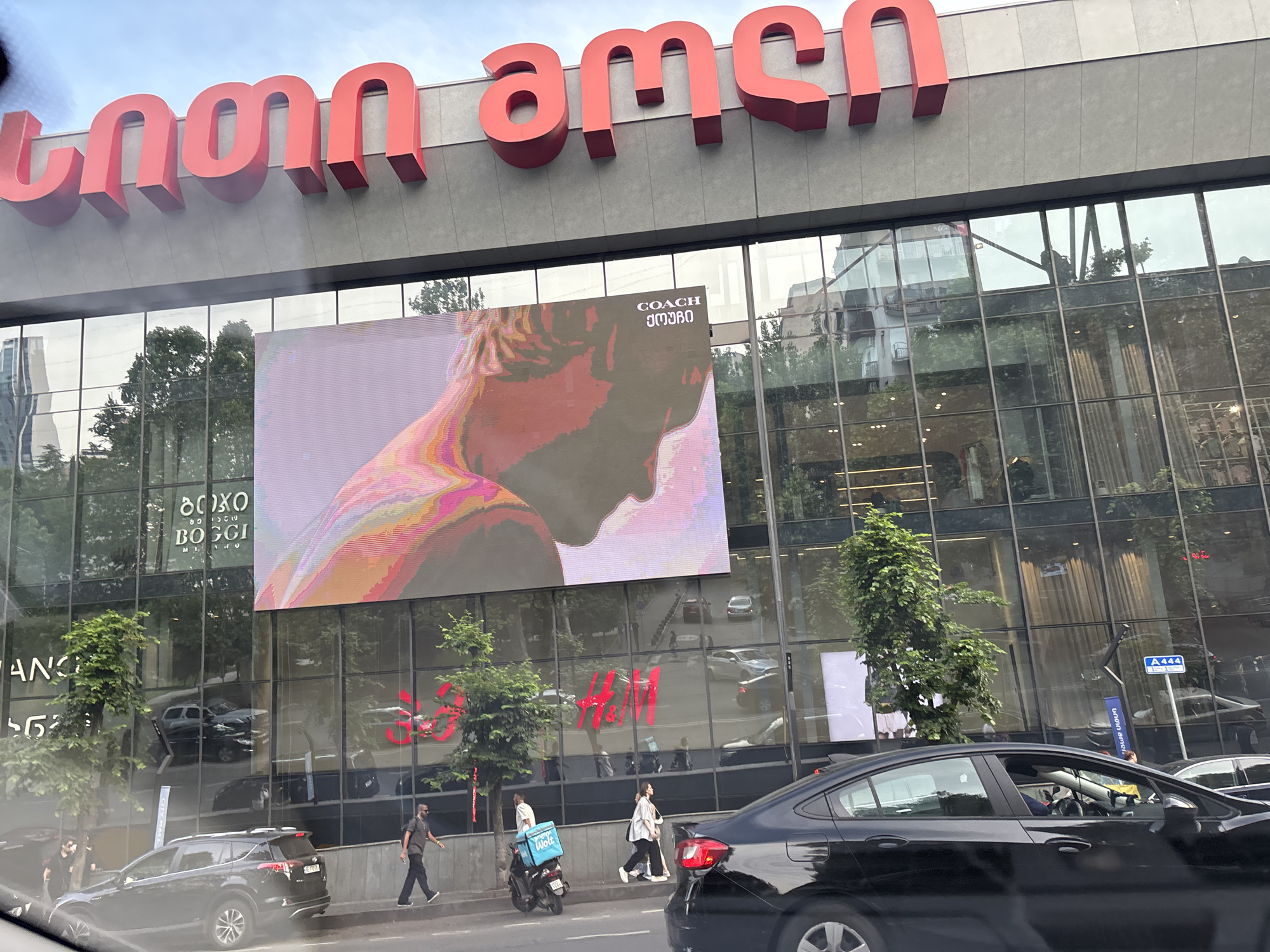 This is a medium-distance, eye-level, outdoor, daytime, slightly angled shot of a modern commercial building in Tbilisi, Georgia. The building features a large, red Georgian script sign on its facade, which reads "თბილისი მოლი" (Tbilisi Mall). Below this, large glass windows reflect the sky and surrounding trees. A large digital screen displays an advertisement, primarily featuring the profile of a person with colorful abstract shapes. The words "COACH" and "georgian" are also visible on the screen. Further down, red H&M logos are visible through the glass. The street in front of the building has several parked and moving cars, including a black SUV and a black sedan in the foreground. Several pedestrians are walking on the sidewalk. One person is walking a dog, another is carrying a blue delivery bag on a scooter, and others are casually strolling. Trees line the sidewalk, and traffic signs are visible. The sky is overcast.