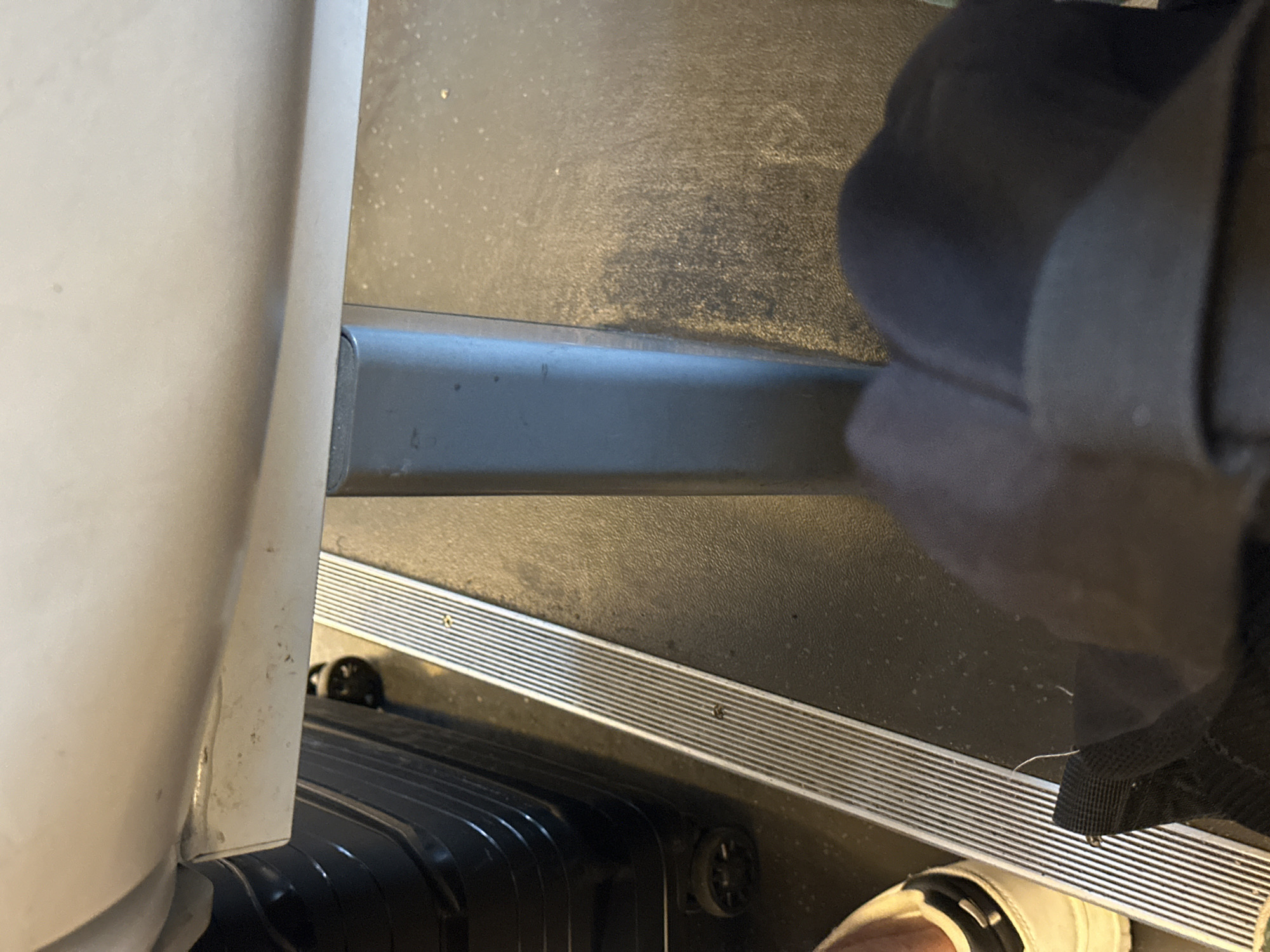 The image shows a close-up, low-angle view of the floor area underneath a light-colored surface, possibly a seat or bench. A dark grey, rectangular support column is visible in the center of the frame. To the left, a black suitcase with ribbed texture and wheels is partially visible. A narrow, metallic strip with parallel lines runs diagonally across the floor. In the lower left, a white shoe with a black stripe and laces is present, and a black bag with a strap is in the foreground, obscuring the lower part of the shoe. The floor appears to be a dark, textured material with some speckling. There are no visible signs of the location being in Uhlstädt-Kirchhasel, Germany, or any specific urban environment.