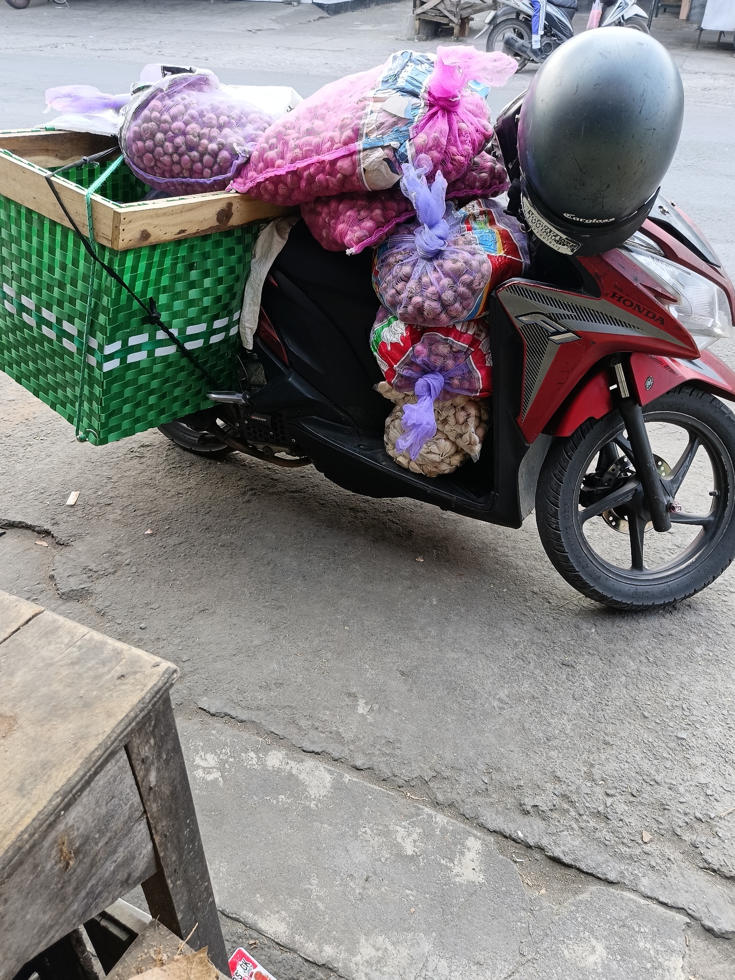 A red Honda motorcycle is parked on a paved street in Sukorini, Indonesia. The motorcycle is loaded with bags of onions. A green woven basket is attached to the back of the motorcycle and contains some of the onions. A black helmet is placed on the handlebars. The street is cracked and worn, with some debris on the ground. A wooden table is visible in the foreground. The background shows other motorcycles and buildings.