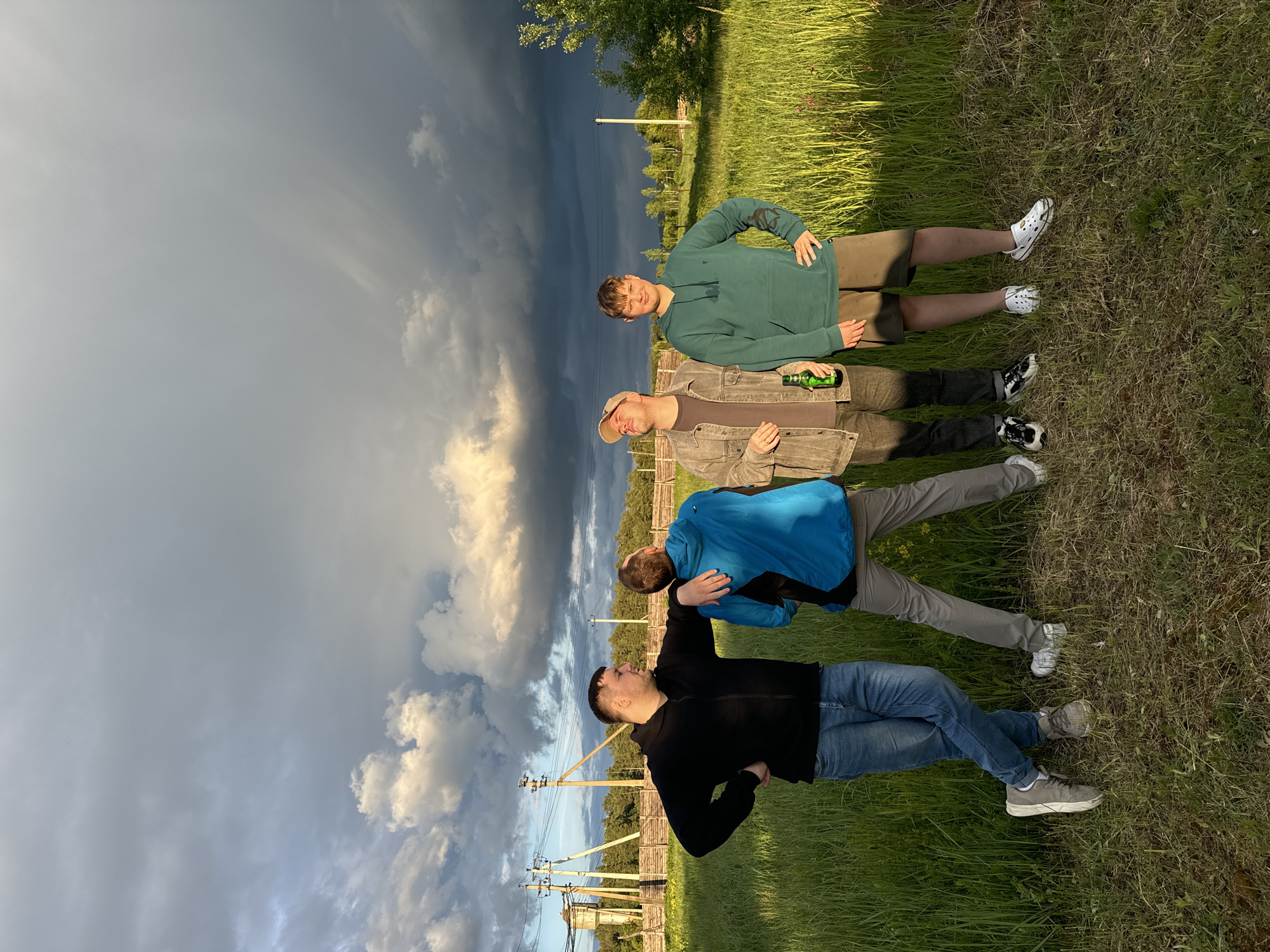 The image depicts four men lying in a grassy field in what appears to be a rural area. In the background, there are utility poles and a line of trees. The sky is dramatic, with a mix of dark, stormy clouds and patches of lighter, sunlit clouds.
From the foreground to the background:
- The first man is wearing a black long-sleeved shirt, blue jeans, and grey sneakers. He is positioned with his arm raised towards the sky and another person behind him.
- The second man is wearing a blue jacket and grey pants, lying on his back.
- The third man is wearing a brown jacket over a dark shirt and dark pants, lying on his back and appears to be holding a green bottle.
- The fourth man is wearing a green hoodie and brown shorts with white clogs.
The grassy field appears to be unkempt, with varying lengths of grass. The overall impression is one of a casual outdoor setting. The location could be Vysokinichi, Russia, based on the request.
