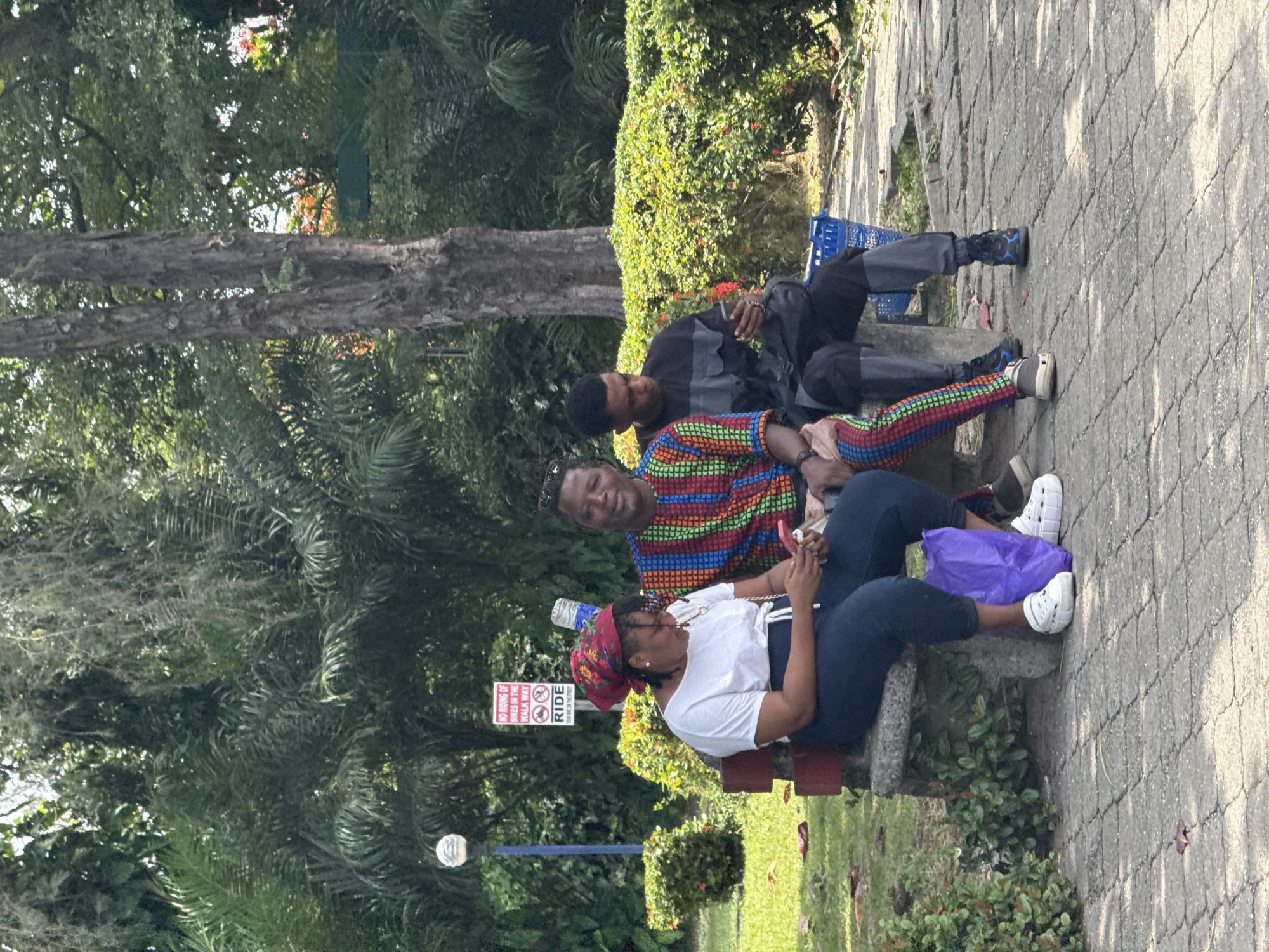 Three people are seated outdoors in what appears to be a park or public green space in Lekki, Nigeria, under bright daylight. The image is oriented vertically but depicts the scene horizontally.
In the foreground, a woman is seated on a concrete bench. She wears a white t-shirt, dark pants, a red patterned headscarf, and white Crocs. She is looking down at a mobile phone in her hands. A purple plastic bag rests near her feet. Next to her on the bench is a man wearing a vibrantly patterned, multi-colored shirt, dark pants, and sunglasses resting on his head. He looks towards the front. A plastic water bottle is on the bench near the woman.
Behind them, further to the left, a third person, a man, is seated on the ground or a lower structure, leaning back. He wears a dark long-sleeved shirt with gray sections and dark pants with dark athletic shoes featuring blue accents. A blue plastic basket or crate is visible near him.
The setting is characterized by lush green vegetation, including tall trees and bushes, suggesting a tropical or subtropical climate. A paved walkway with a patterned surface is visible. The bright sunlight casts distinct shadows, indicating a clear, sunny day.
A red and white sign on a pole in the middle ground displays text: "NO RIDING OF BIKES IN THE WALK WAY" with a crossed-out bicycle icon, and "RIDE YOUR BIKE ON THE STREET" with a plain bicycle icon. A white street lamp fixture is also visible in the left foreground.