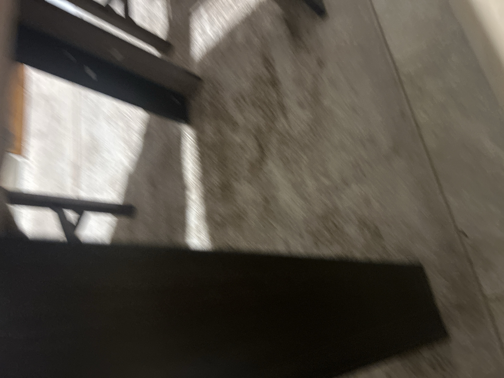 Location: Unknown, Unknown.
The image presents an interior scene, primarily depicting a section of a floor and the lower portion of furniture. A dark, vertically oriented rectangular object, consistent with a furniture leg or structural support, dominates the left side of the frame, extending from the bottom edge to approximately three-quarters up. The floor surface appears to be a light-colored, textured material, possibly a rug or carpet, with areas of varying shade and detail. Numerous dark shadows are cast across this floor, indicating directional illumination from an unseen light source. One distinct shadow in the upper-right quadrant suggests the presence of another vertical furniture element outside the immediate focus. Multiple linear and irregular shadow patterns are visible across the floor, intermingling with brighter, illuminated sections. Faint outlines of additional dark, linear structures are discernible in the background. The image exhibits significant motion blur, particularly affecting the texture of the floor and the clarity of shadow edges. No individuals, animals, or specific actions are observable.