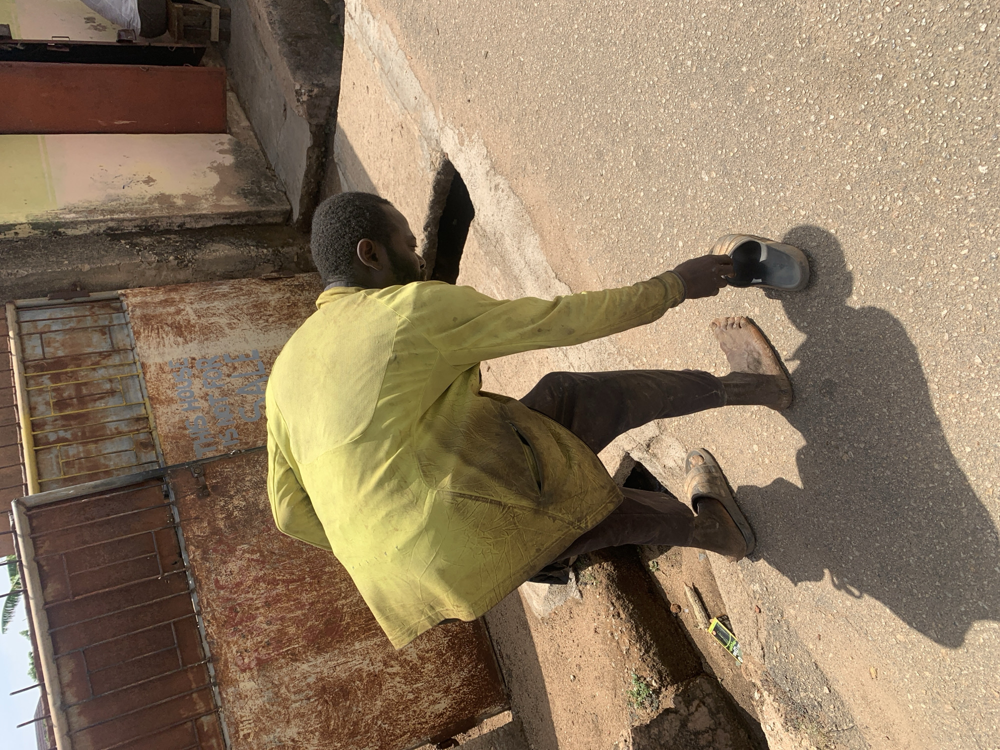 A man in a light green jacket and dark trousers stands on a paved surface, potentially a street, with a sidewalk on one side. He is barefoot and holding a sandal in his right hand. The man is near a rusty metal gate or door with a barred window. The background features concrete structures and vegetation. The scene is lit by sunlight, casting shadows of the man and the sandal. There is a pack of cigarettes or another item on the ground near his foot. The location is Jos, Nigeria.