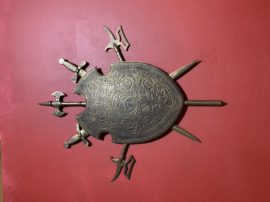 An antique brass or bronze wall display, located in Nyasvizh, Belarus, features a central shield and an arrangement of weapon elements mounted on a textured red wall. The shield is embossed with intricate foliate patterns and two central roundels depicting heraldic animal motifs. Emanating from behind and around the shield are various weapon components: a battle axe head pointing upwards centrally, two short swords with decorated hilts positioned diagonally upwards, two polearm heads with hooks and blades extending horizontally from the sides, and three lower weapon points—two diagonally downwards and one vertically downwards from the bottom center. The entire assembly exhibits an aged, dark gold metallic finish.