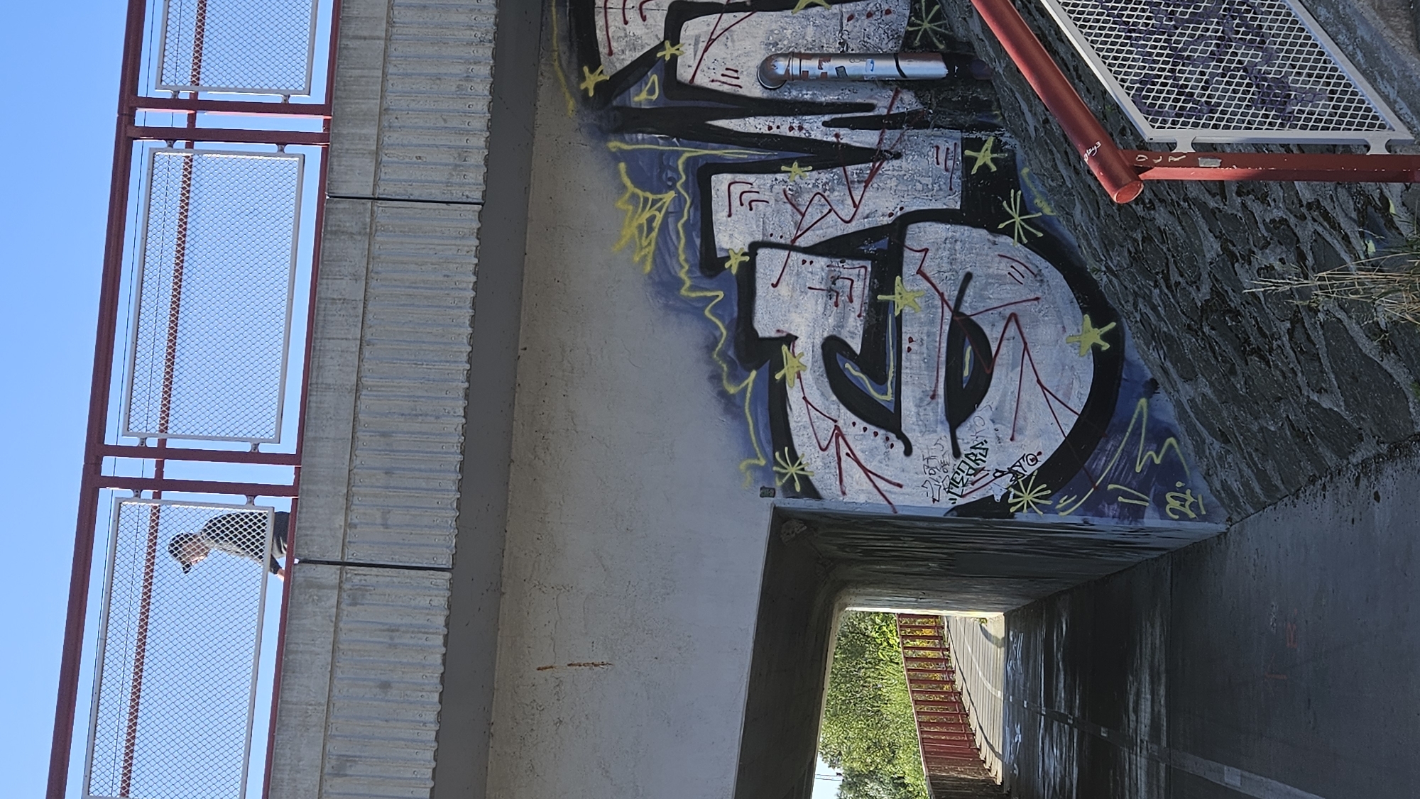A person wearing a black baseball cap and a dark jacket walks on a pedestrian bridge. The bridge has a red metal railing with a wire mesh infill. The bridge is constructed of concrete and runs over a pathway. On the concrete wall beneath the bridge, there is graffiti featuring abstract shapes and lettering in black, white, yellow, and red. The pathway appears to be a bike path or walkway with a red railing alongside it. The background shows a clear blue sky and some greenery. The overall scene appears to be an urban environment.