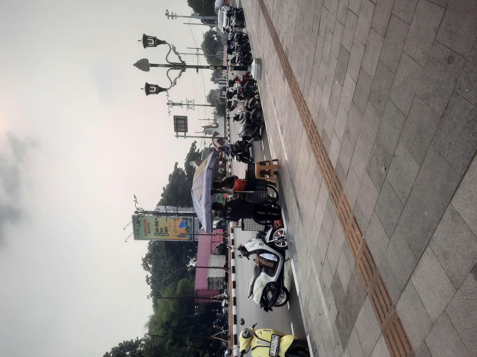 The image depicts a street scene in Sukaraja, Indonesia, under an overcast sky. A row of parked motorcycles lines the street, with a yellow scooter in the foreground on the left. Adjacent to the parked motorcycles, a street vendor operates under a blue and white umbrella, serving customers. The vendor's cart is visible, with items displayed. Two individuals are interacting at the vendor's stall. Beyond the vendor and motorcycles, the street is busy with traffic, including cars and more motorcycles. The background features buildings, trees, and a billboard advertising "Coming Soon". The sidewalk is paved with grey tiles, and a tactile paving strip runs along its edge. The overall environment suggests a typical urban setting with commercial activity.