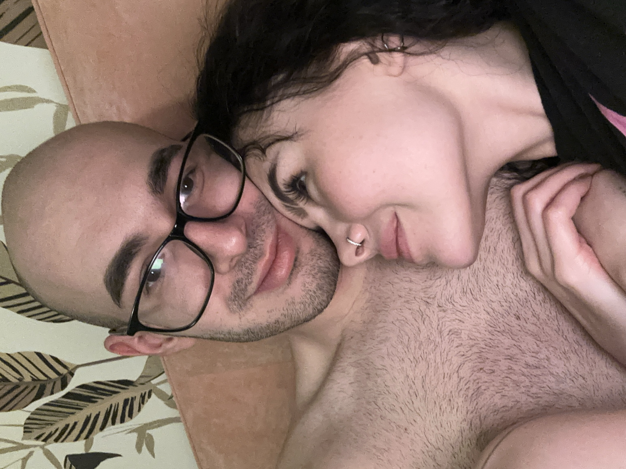 A close-up selfie shows a couple lying down, likely indoors. A bald man wearing black-rimmed glasses is on his back, looking at the camera. A woman with dark, curly hair is resting her head on his chest, her face close to his. She has a nose ring. The man's chest is visible and has chest hair. The background is a floral-patterned surface and a peach-colored cushion or headboard. The location cannot be determined as Kazan, Russia from the image alone.