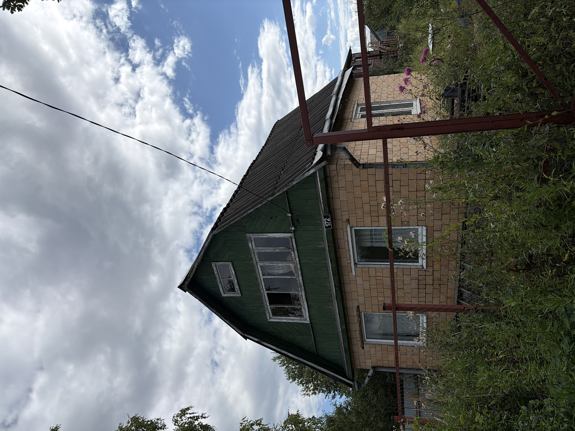 The image depicts a two-story house in the SNT Keramik (Belaya) area, Russia. The house has a dark, sloped roof and a green-painted upper level with several windows. The lower level is constructed of light brown bricks. A red metal fence encloses the property, which is surrounded by overgrown vegetation and some flowering plants. The sky is partly cloudy with a mix of gray and blue. A power line runs diagonally across the upper portion of the image. The house appears to be residential, likely a dacha or summer house, and the overall scene suggests a rural or semi-rural setting.